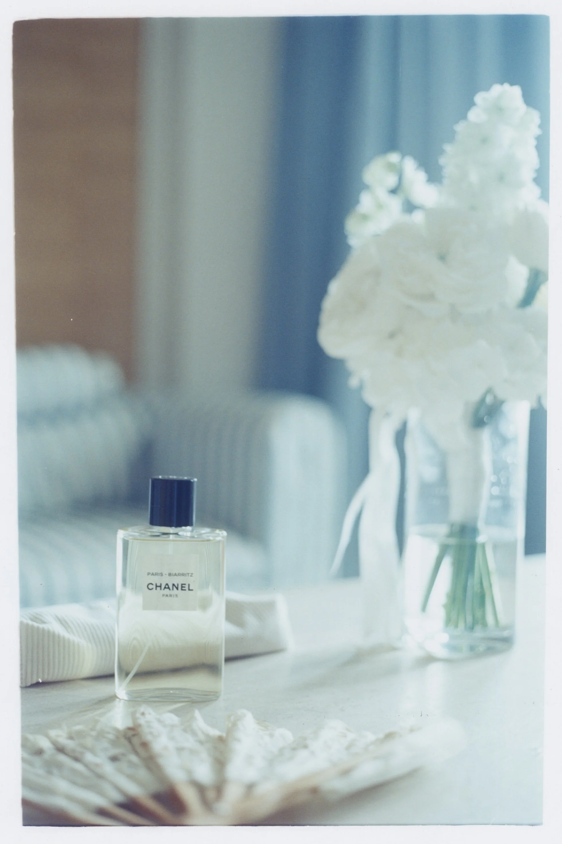 Still-life photo of a bride's accessories on her wedding day. The chosen film increases that blue ambiance and softens the colors, as it's a film that's used for cinema. We can see a bottle of Chanel perfume, a white flower bouquet and a fan. 
Photo 