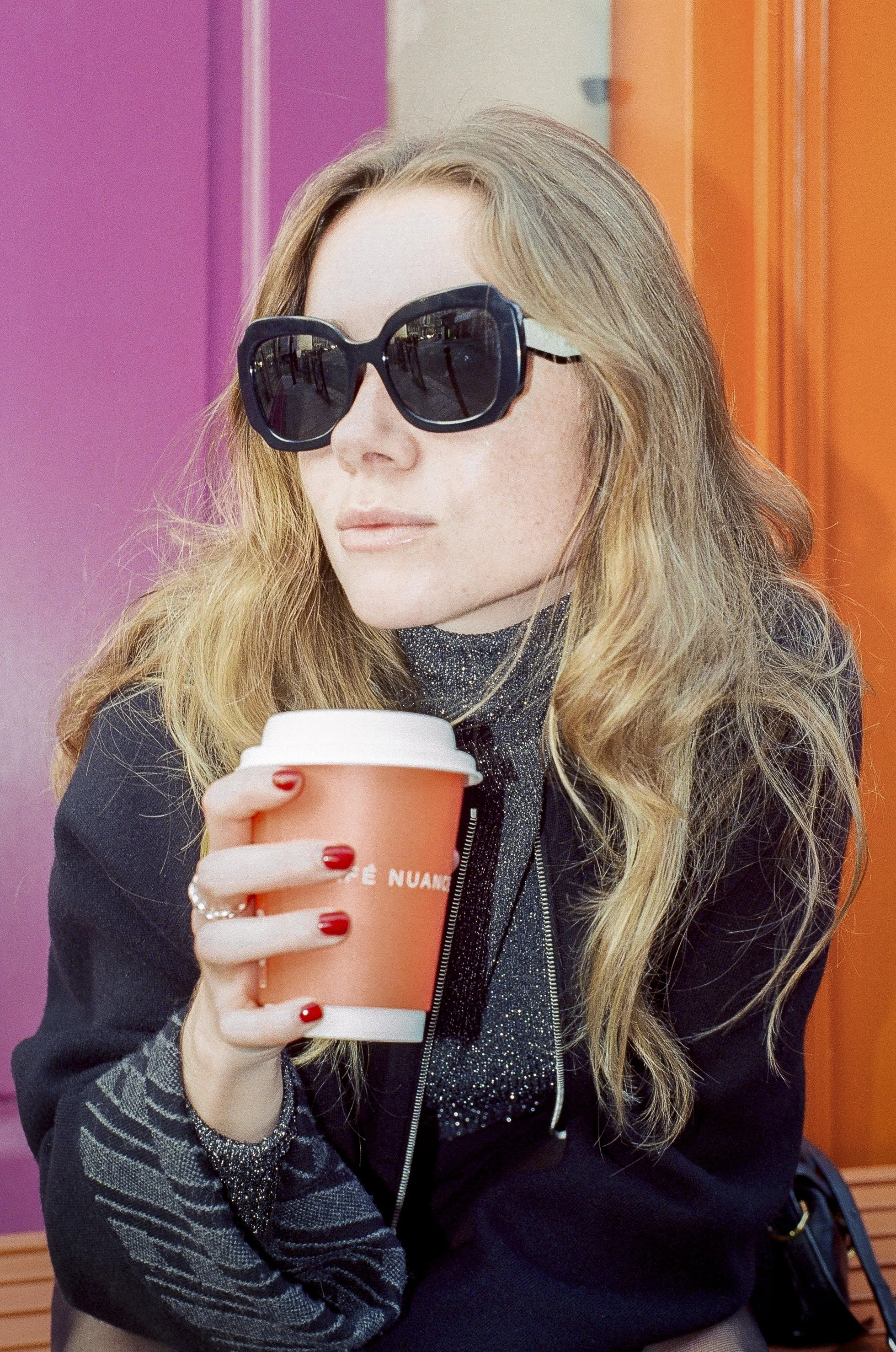 Femme avec lunettes de soleil noire et cheveux blonds, tenant un café dans une tasse orange dans un décor coloré.