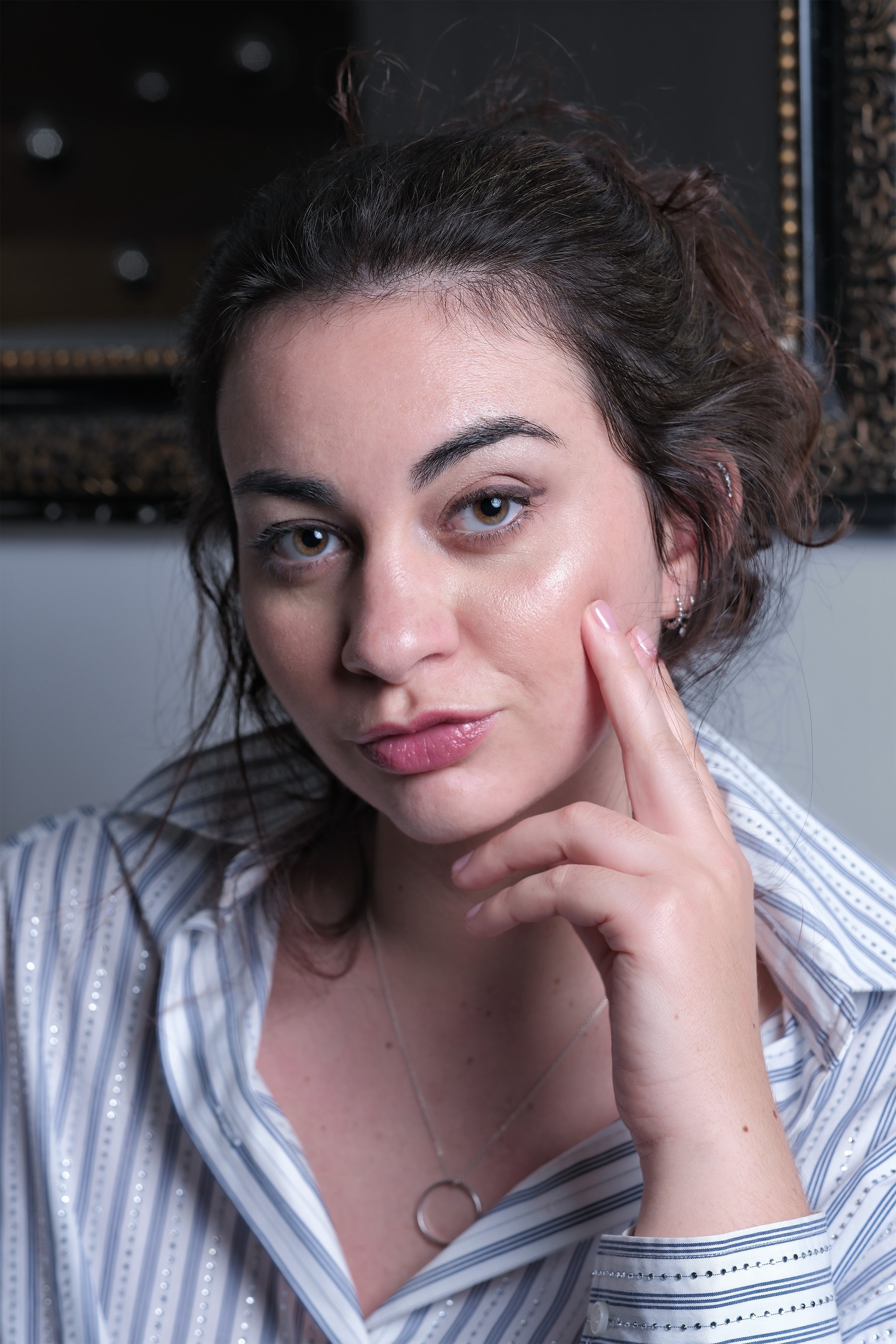 Un portrait d'une femme aux cheveux bouclés marron clair, portant une chemise à rayures bleues et blanches, avec des boucles d'oreilles et une chaîne fine, regardant vers l'objectif et touchant son visage avec son index.