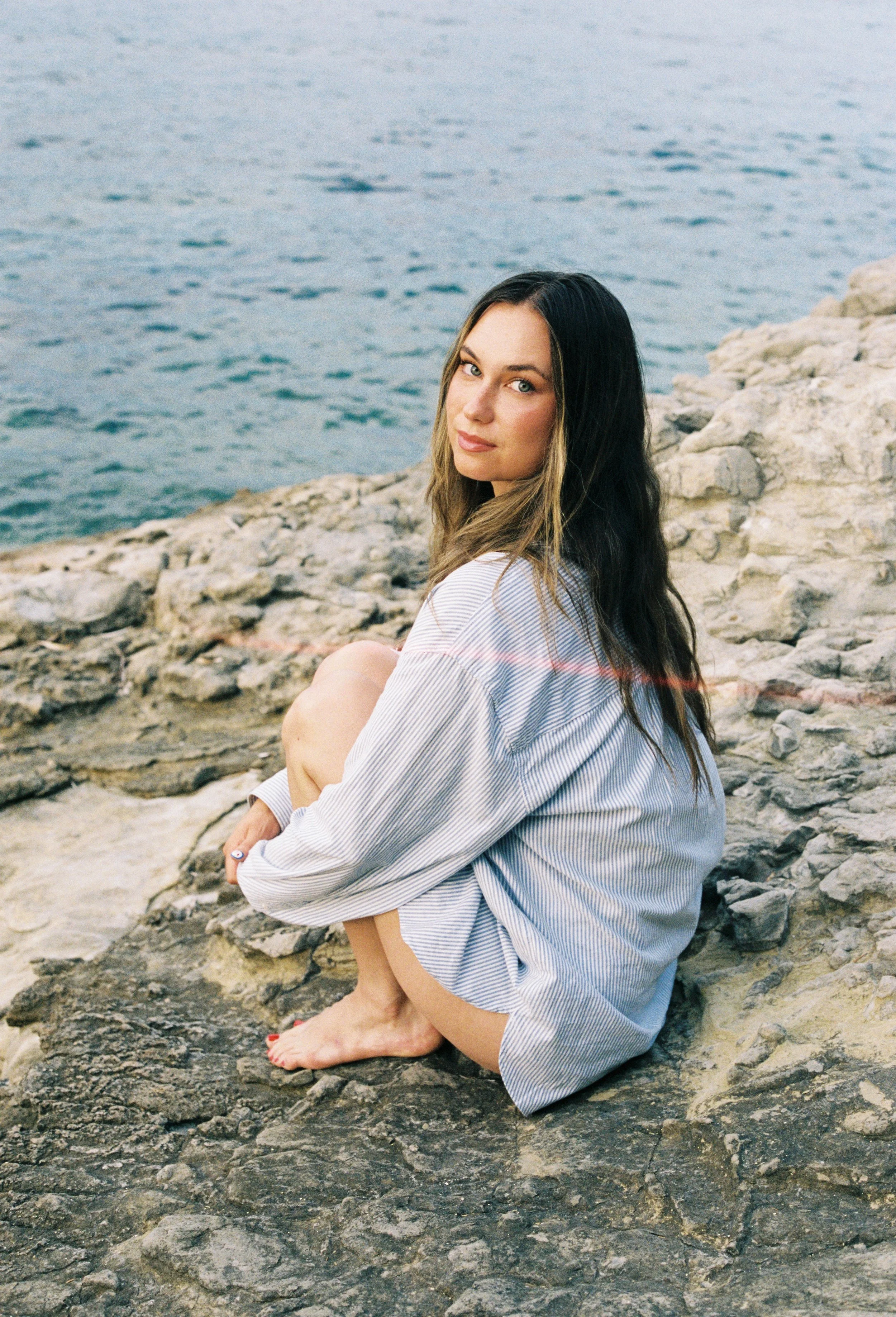Photo of a woman showing her entire body, only wearing a bathing suit and a long shirt. She's sitting on the rocks by the beach. Cold ambiance. 
Photo taken in Saint-Jean-Cap-Ferrat (South of France, French Riviera) on a rock beach. 
Film photo, 35mm