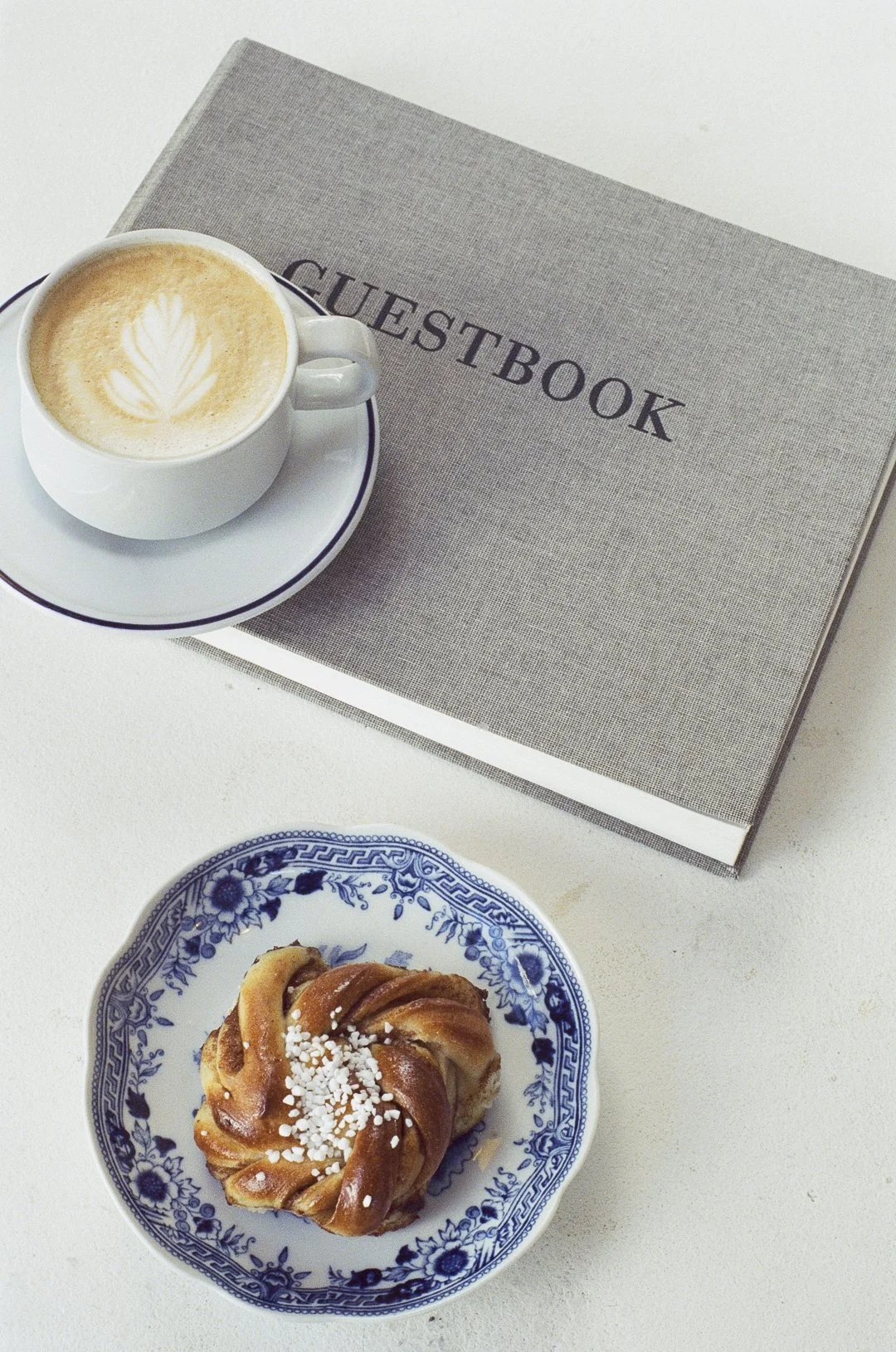 Une tasse de café avec un dessin de latte art, un livre intitulé 'Guestbook' et une viennoiserie sur une assiette en porcelaine.