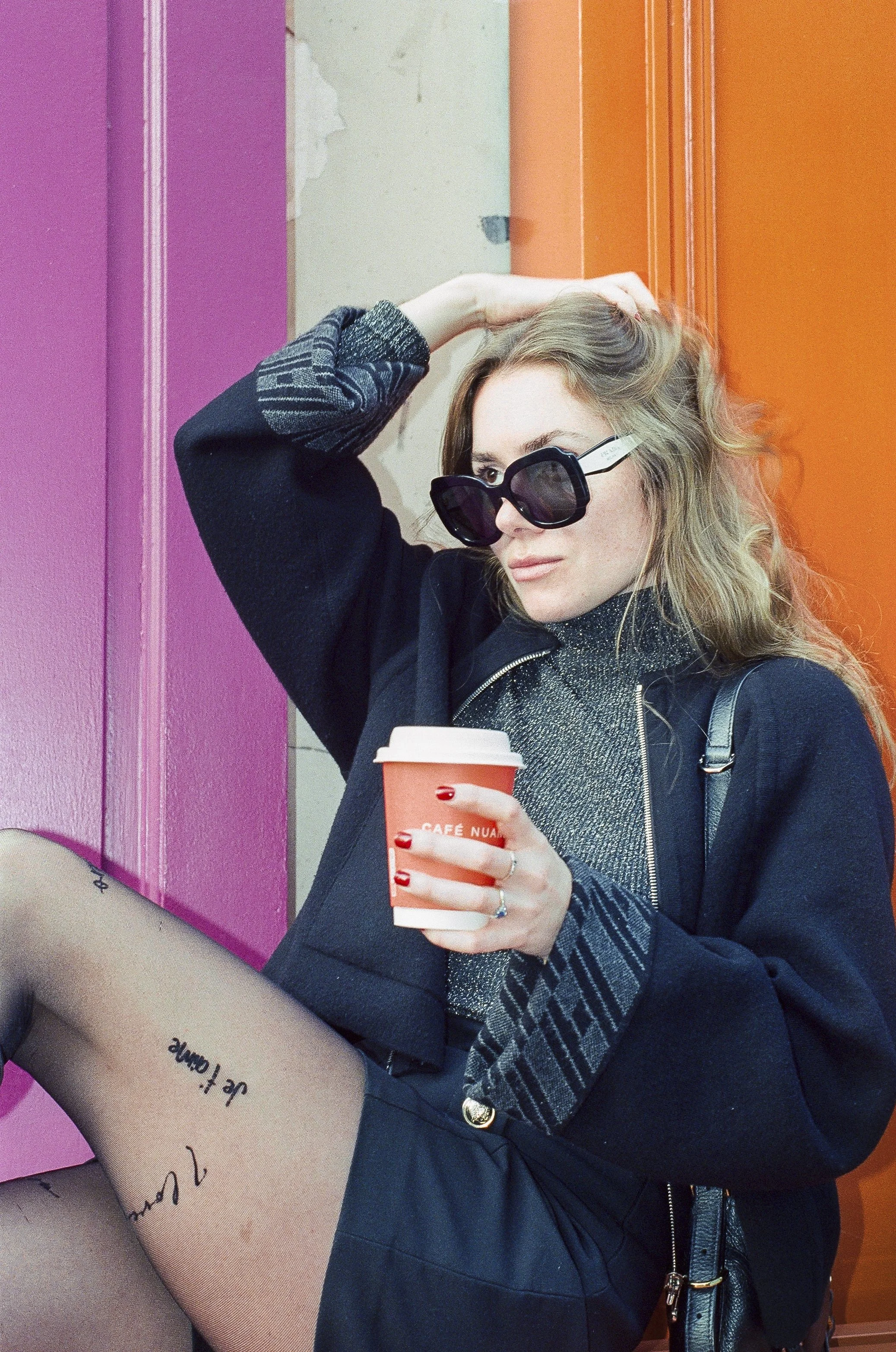 Une femme portant des lunettes de soleil, tenant une tasse de café, assise contre un mur rose et orange.