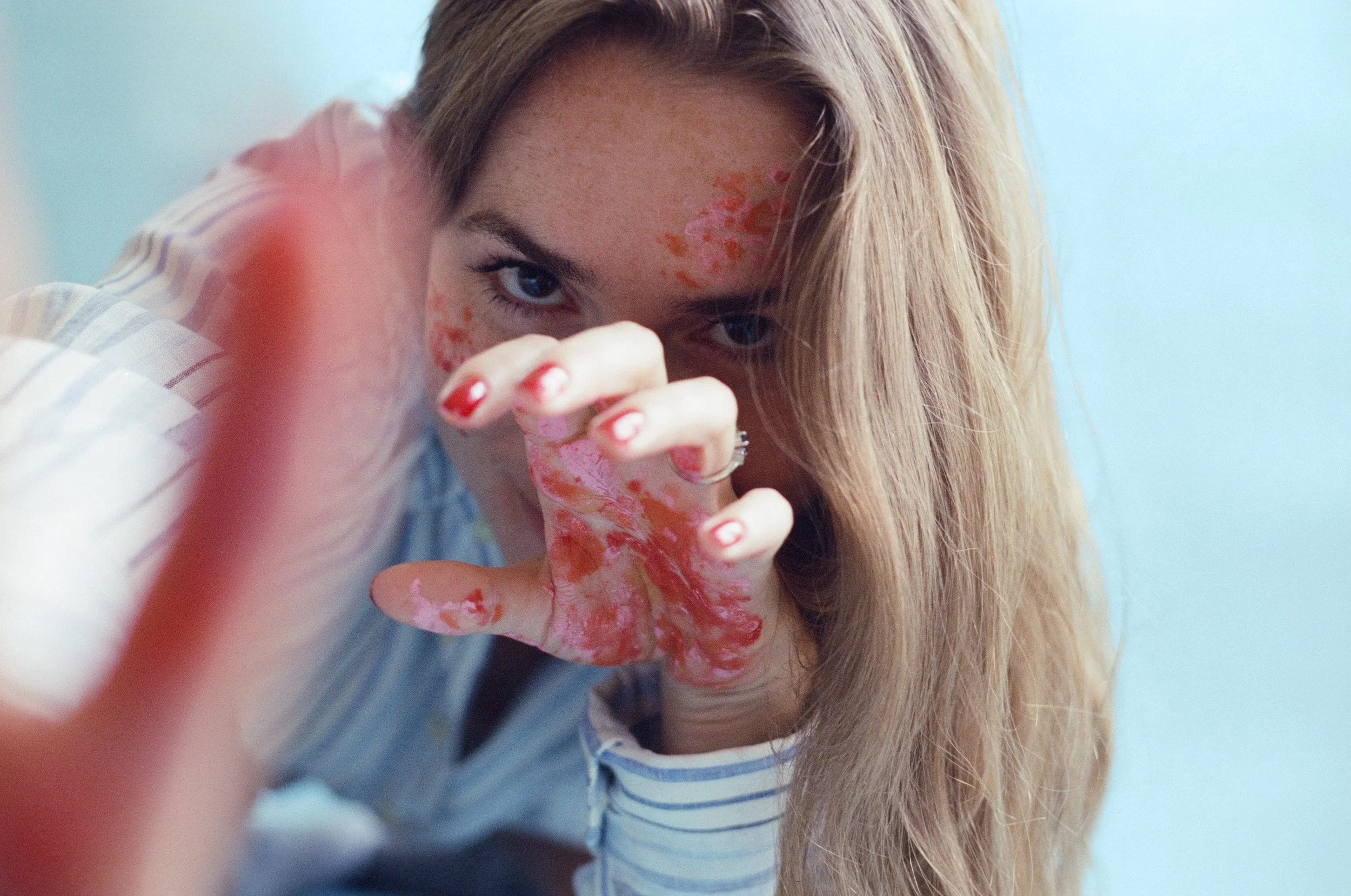 Portrait of a woman showing her upper body. She's wearing a shirt and has paint stains on her face and her hands. The photographer played with perspective by using a small aperture, giving that result of a blurry finger on the first plan and a sharp 