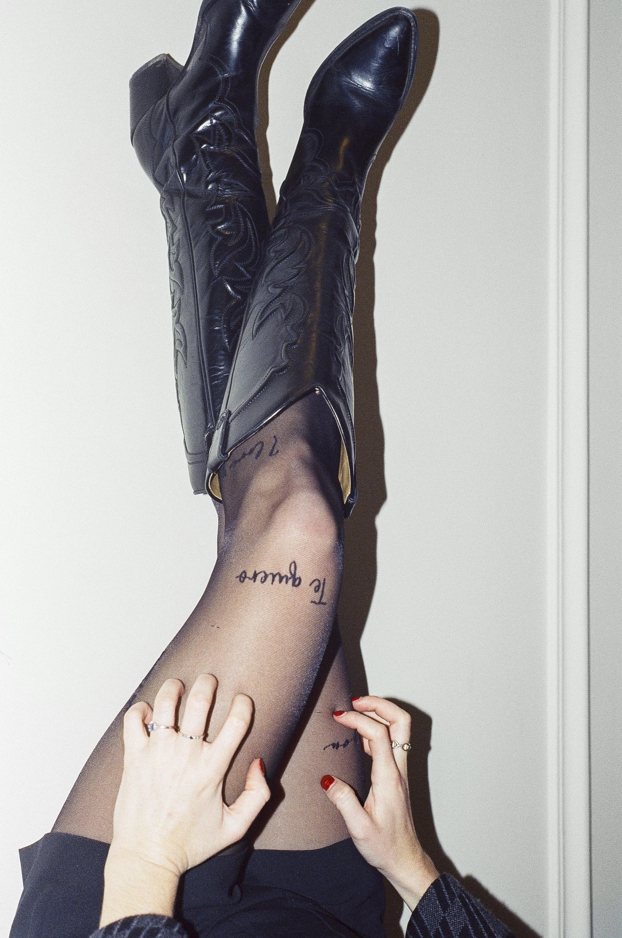 Jambes féminines vêtues de bas en nylon noir, portant des bottes en cuir noir, posées contre un mur blanc, avec des mains ajustant le nylon.