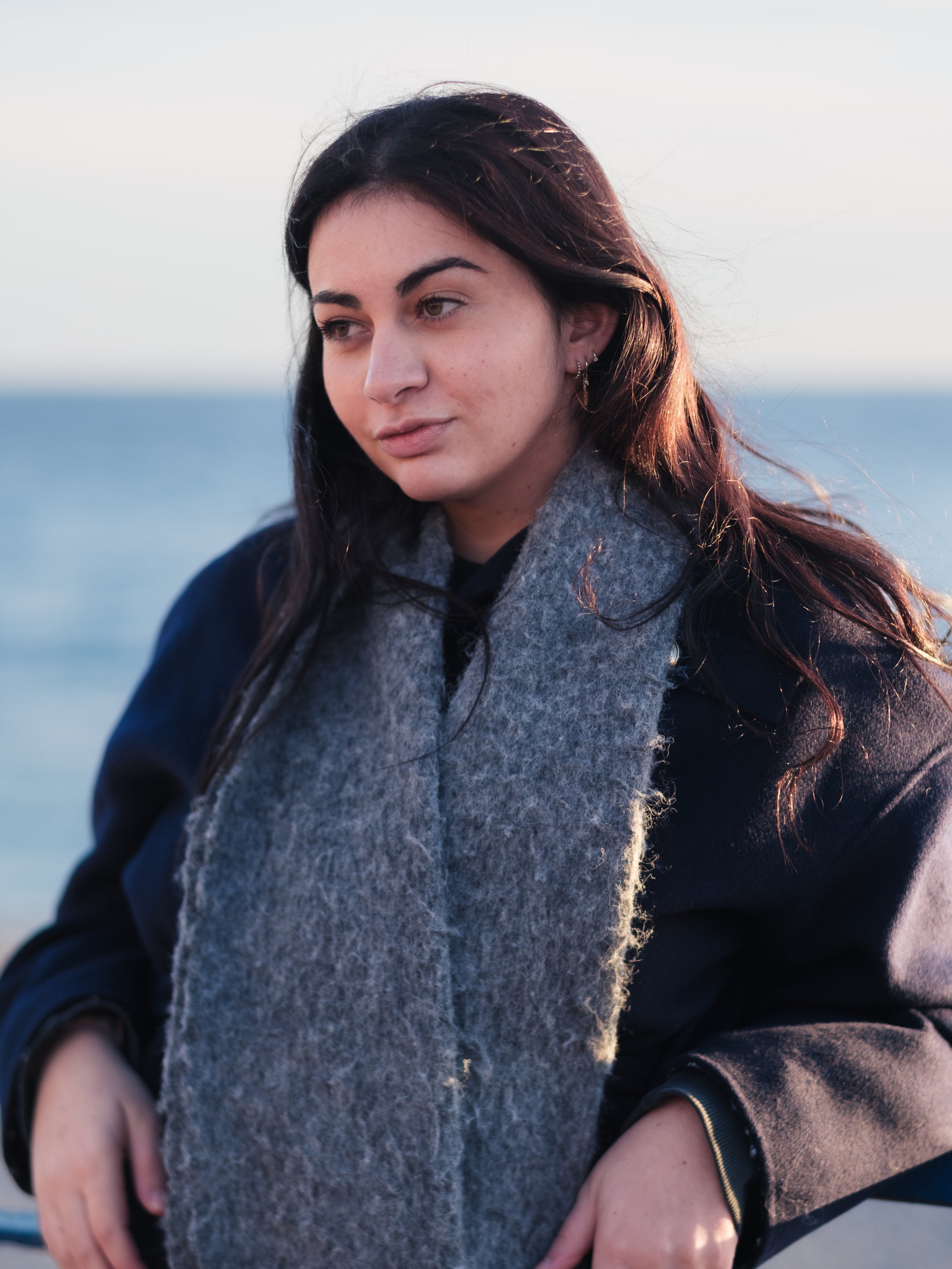 Une jeune femme avec des cheveux bruns foncés, portant un manteau noir et une écharpe grise, est près de la mer en fin d'après-midi ou au coucher du soleil.