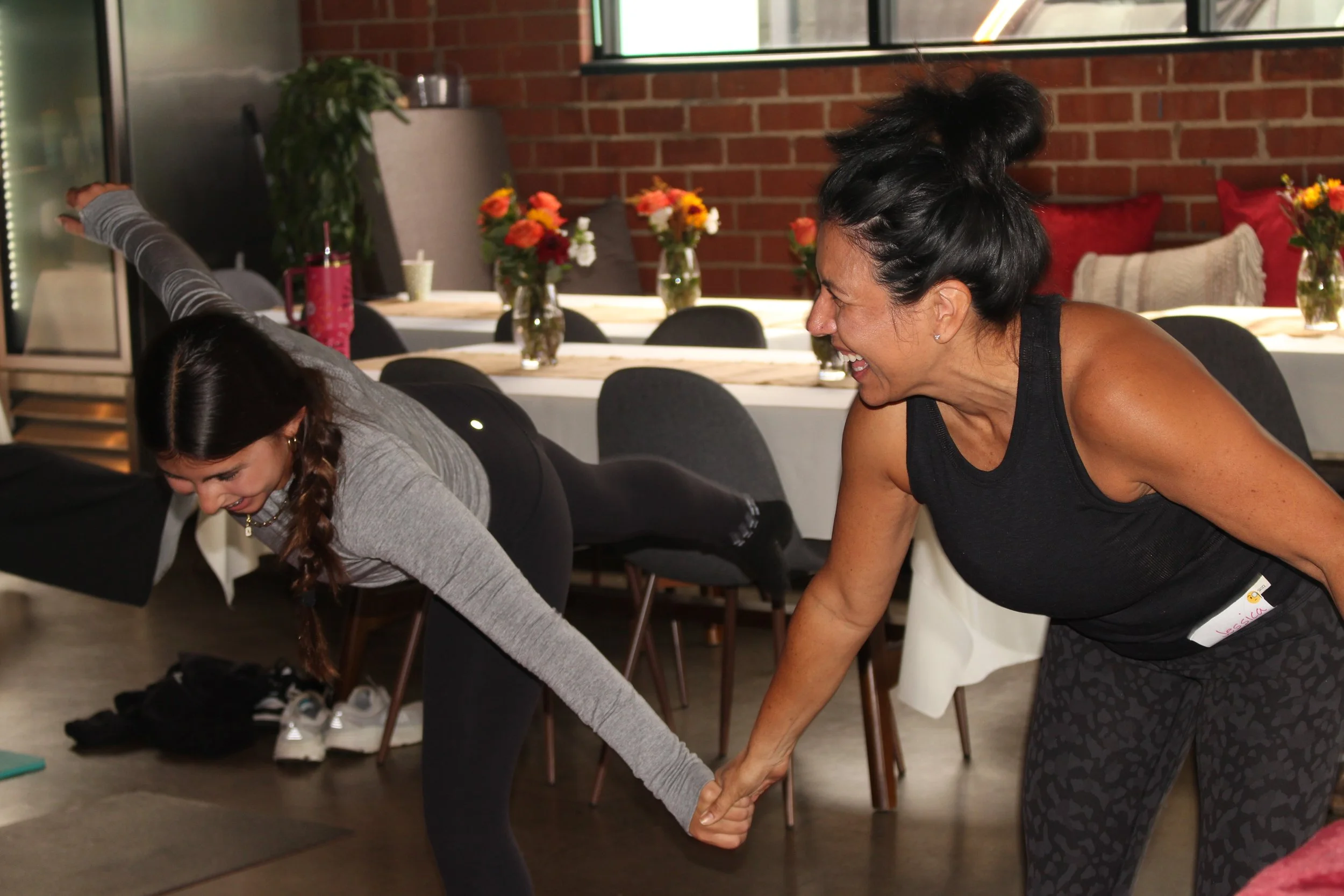 Two women smiling and holding hands during a workout in a room with a table, chairs, and flower vases.