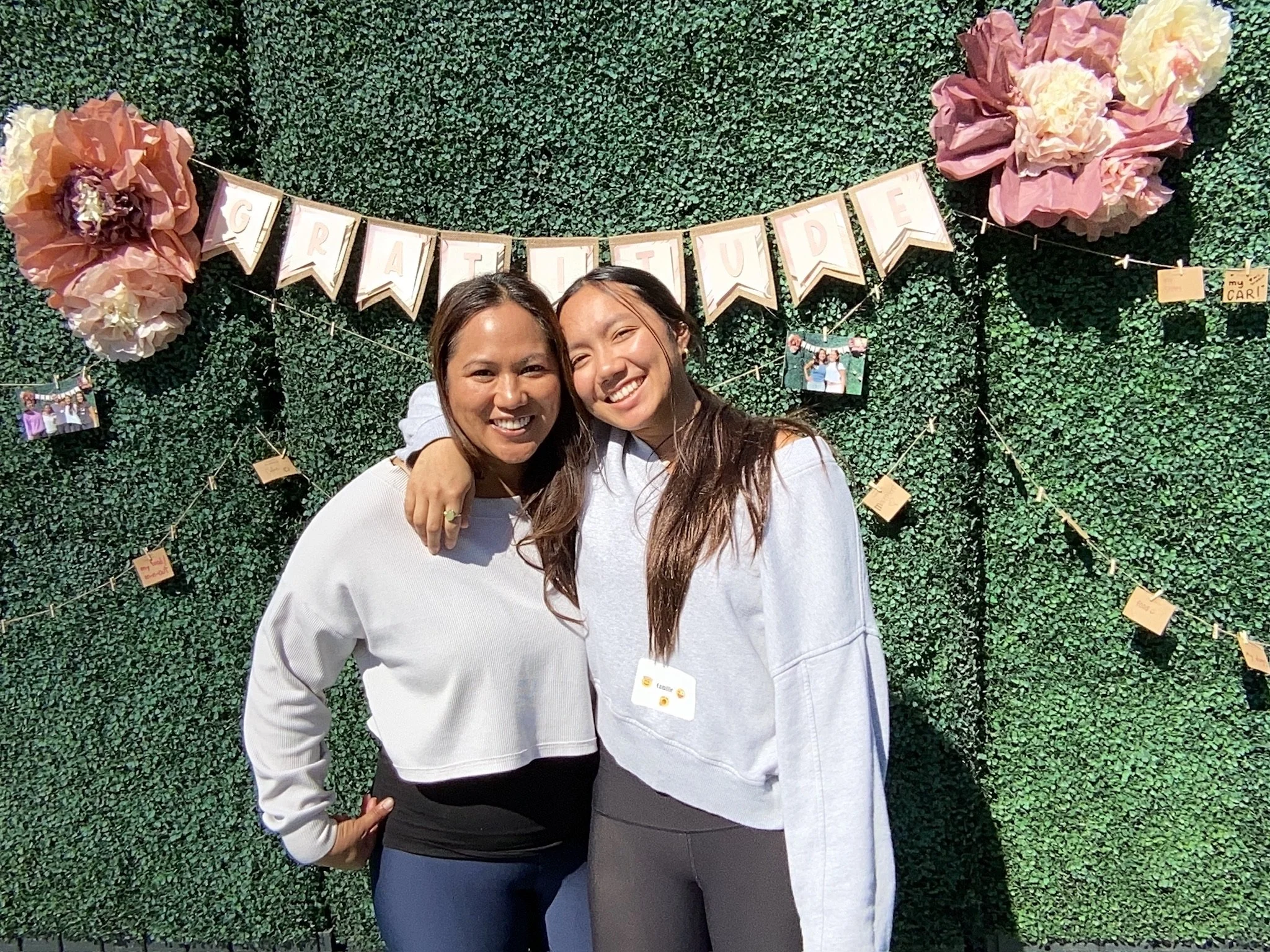 Two women smiling and embracing each other in front of a green hedge wall decorated with a 'Congratulations' banner, large pink and cream paper flowers, and hanging photos.