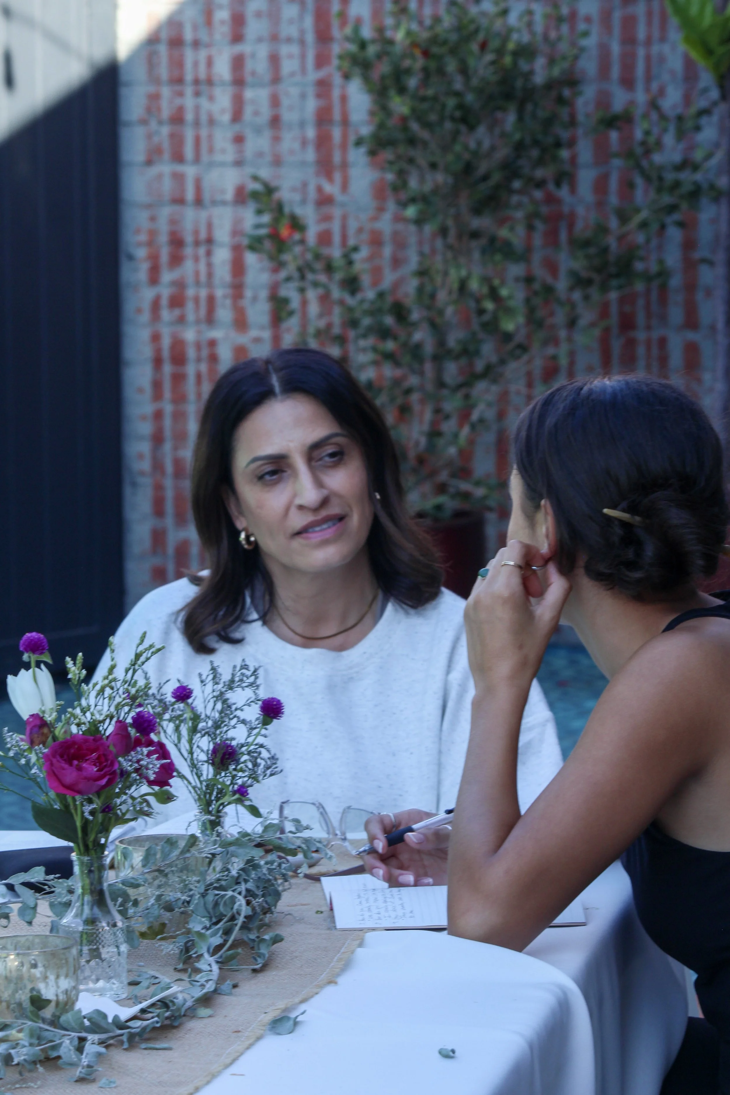 Two women are sitting at a table outdoors, engaged in a conversation. One woman, with dark hair and wearing a white top, appears to be listening or speaking thoughtfully. The other woman, with her back to the camera, has dark hair styled in a bun, and is wearing a black top. There is a flower arrangement with pink, purple, and white flowers on the table, along with a glass of water, notes, and a pen. The background features a brick wall, a potted plant, and part of a black fence.