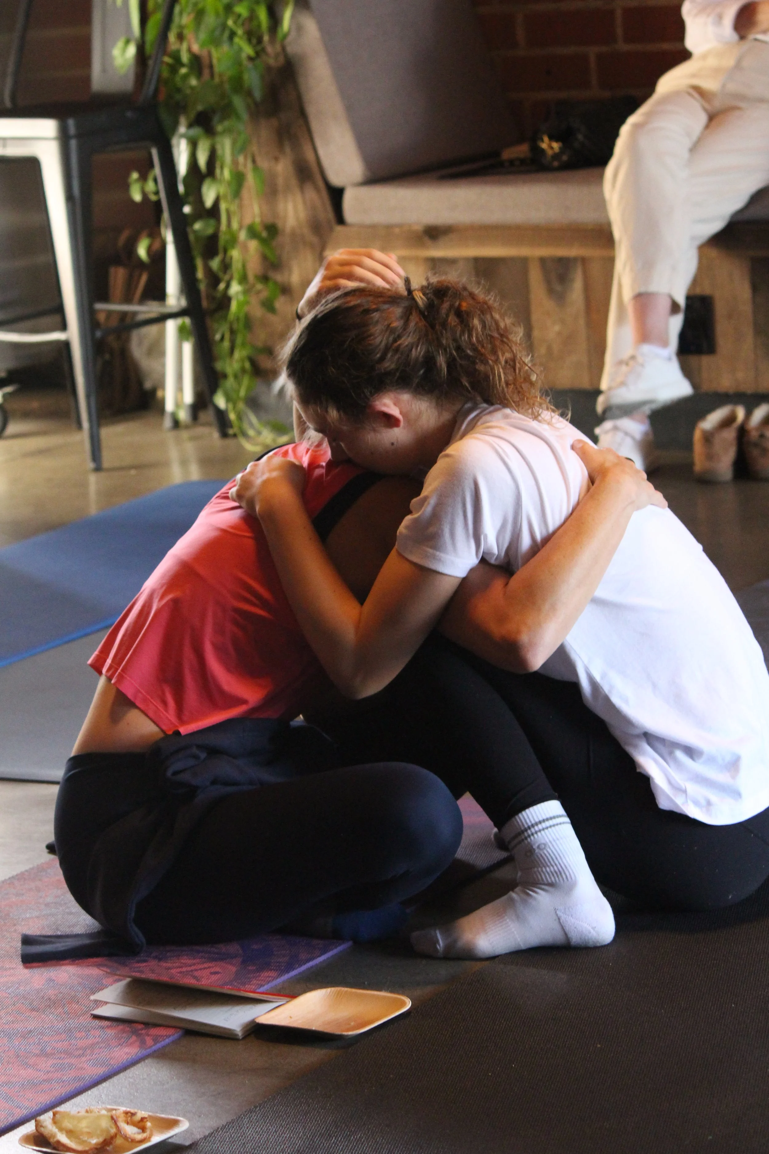 A mom and daughter embracing and comforting each other on yoga mats, in a cozy indoor setting, with a third person sitting in the background. The scene conveys support and emotional connection.