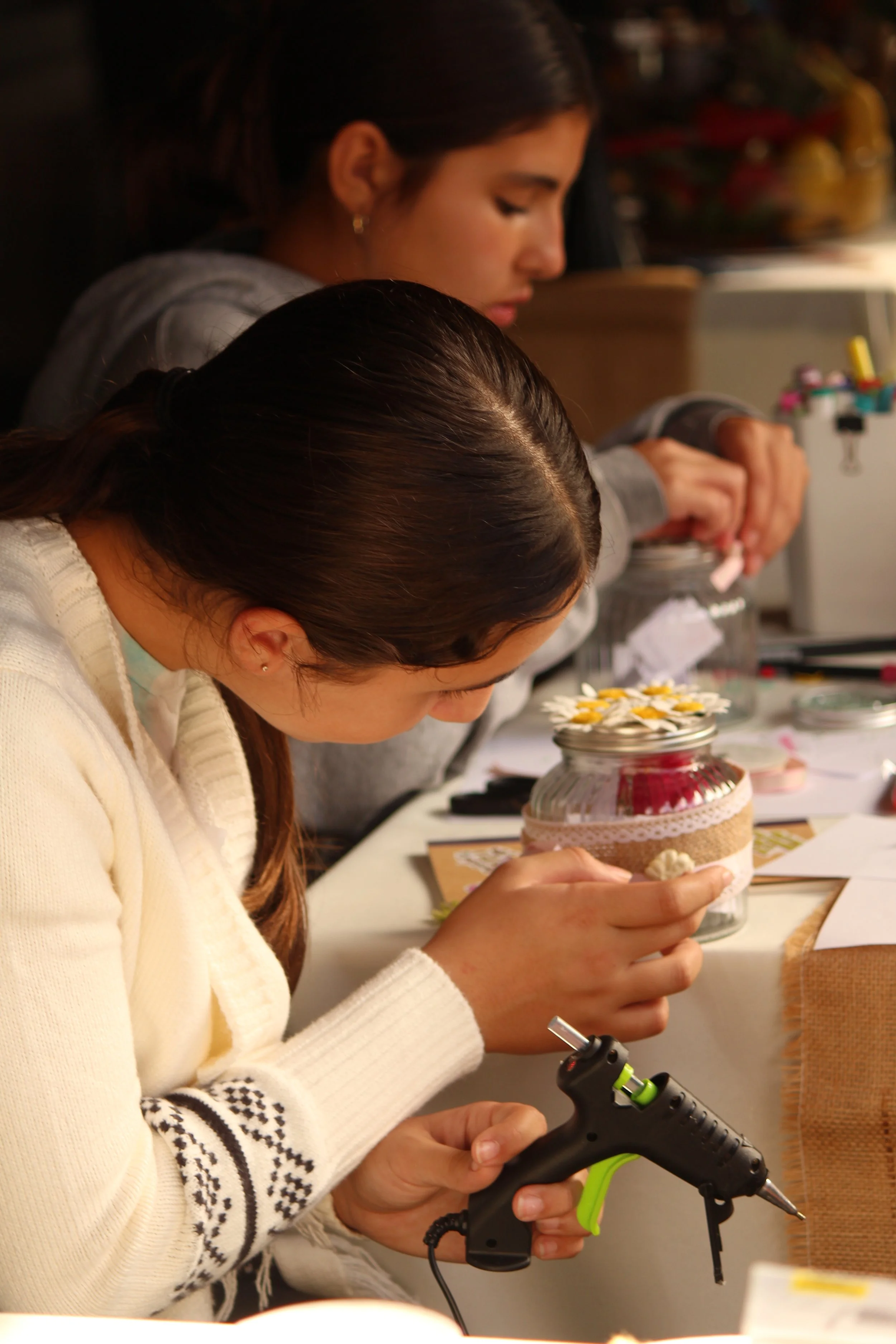 Two young women working on craft projects at a table, one using a hot glue gun, the other decorating a jar with flowers and ribbon.