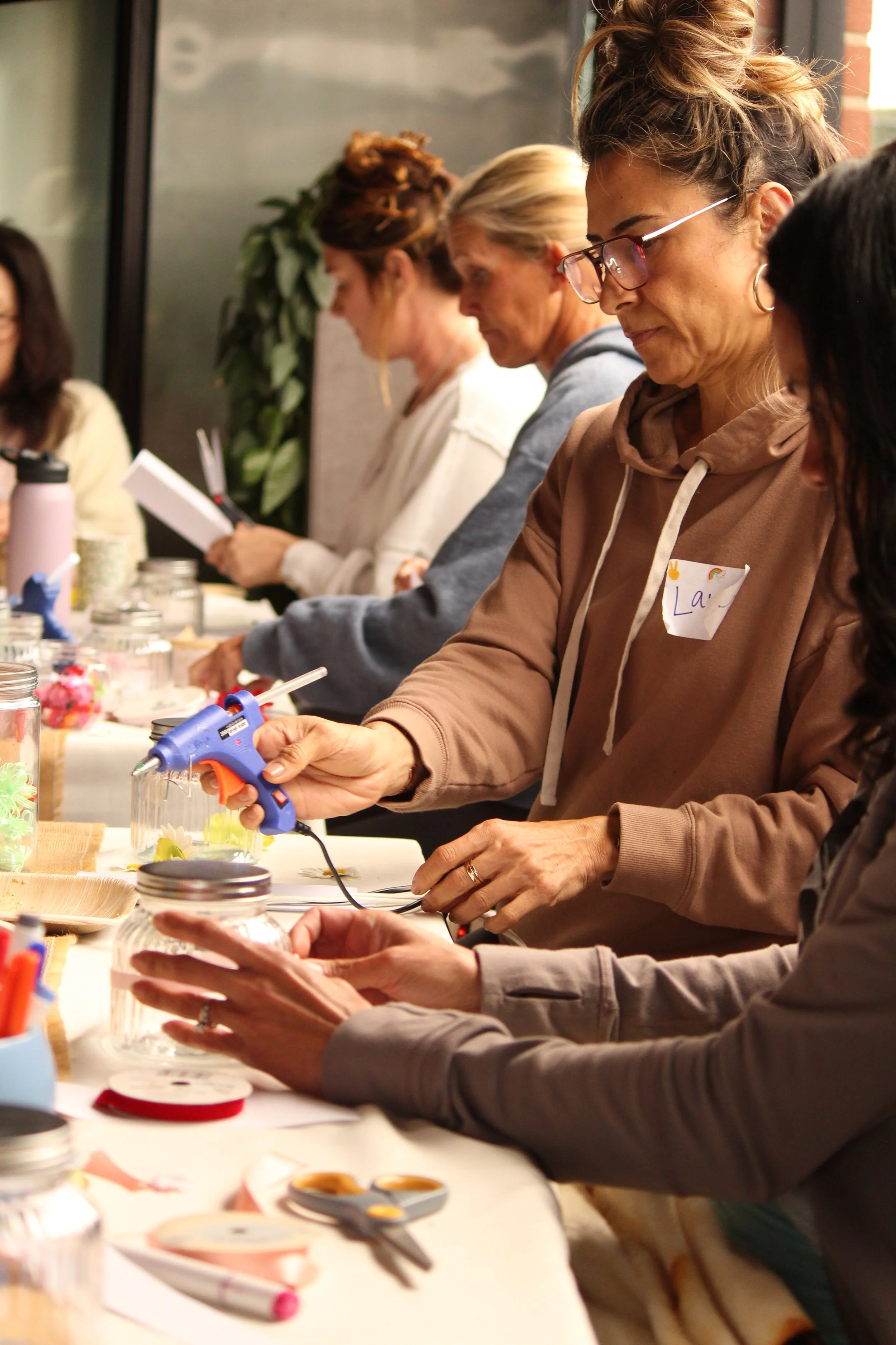 People gathered around a table engaging in arts and crafts activities, using tools like glue guns, scissors, and jars filled with craft supplies.