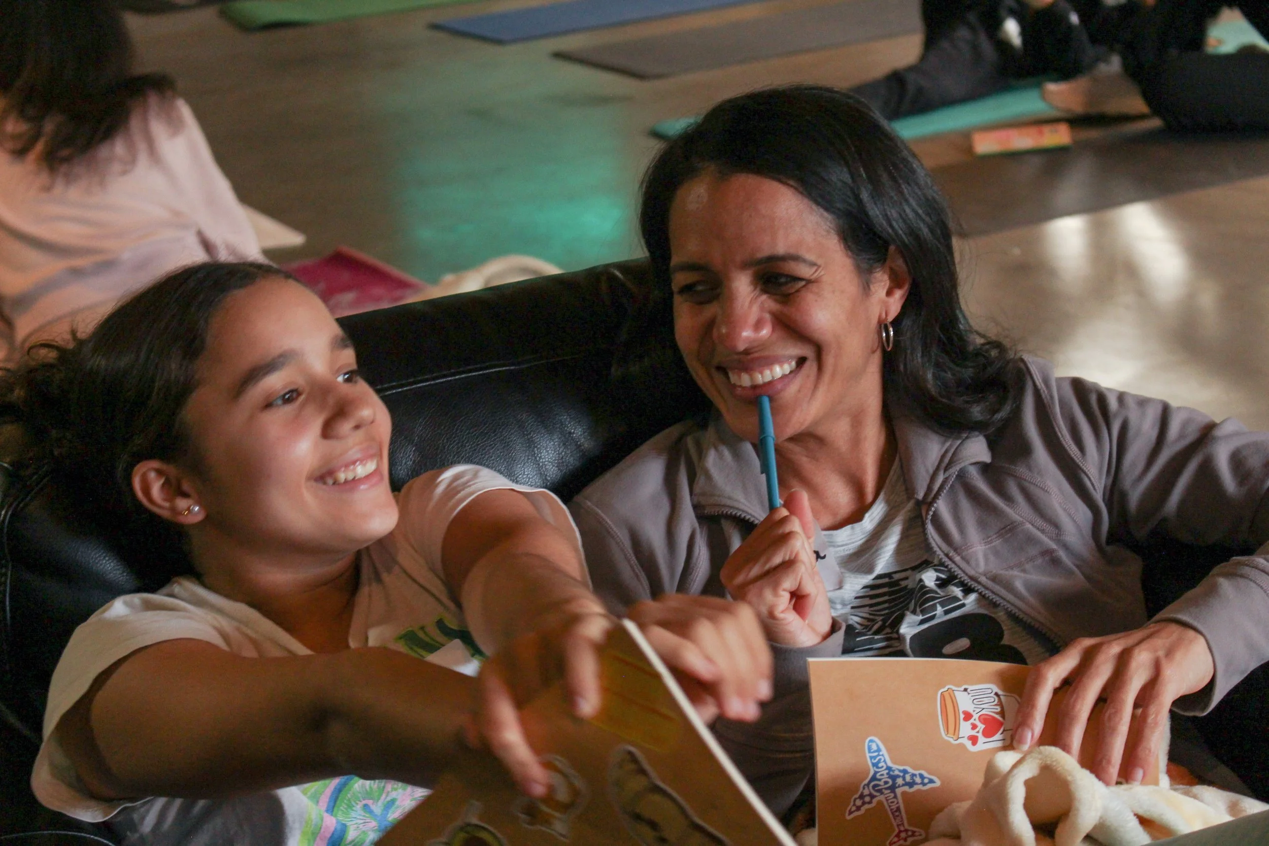 A mom and daughter sitting together on a couch are smiling and enjoying each other's company. In the background, there are other people lying on yoga mats, indicating a relaxed group setting.