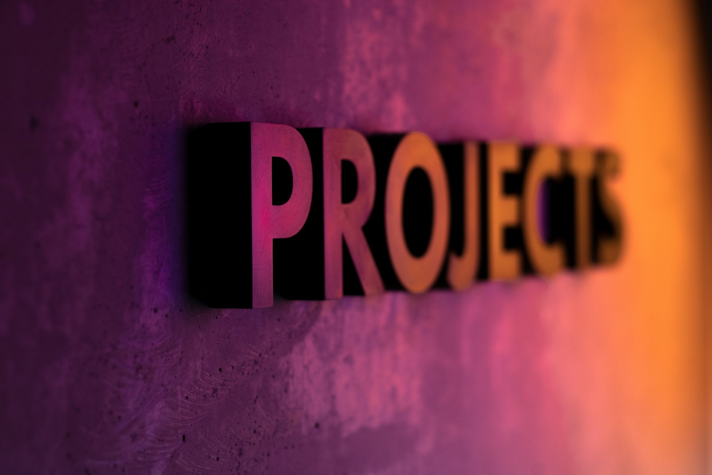 Nahaufnahme von einem 3D-basierten Projektetikett mit den Buchstaben "PROJECT" in pinker Schrift auf einer glatten Oberfläche, beleuchtet mit buntem Lichteffekt in Pink und Orange.