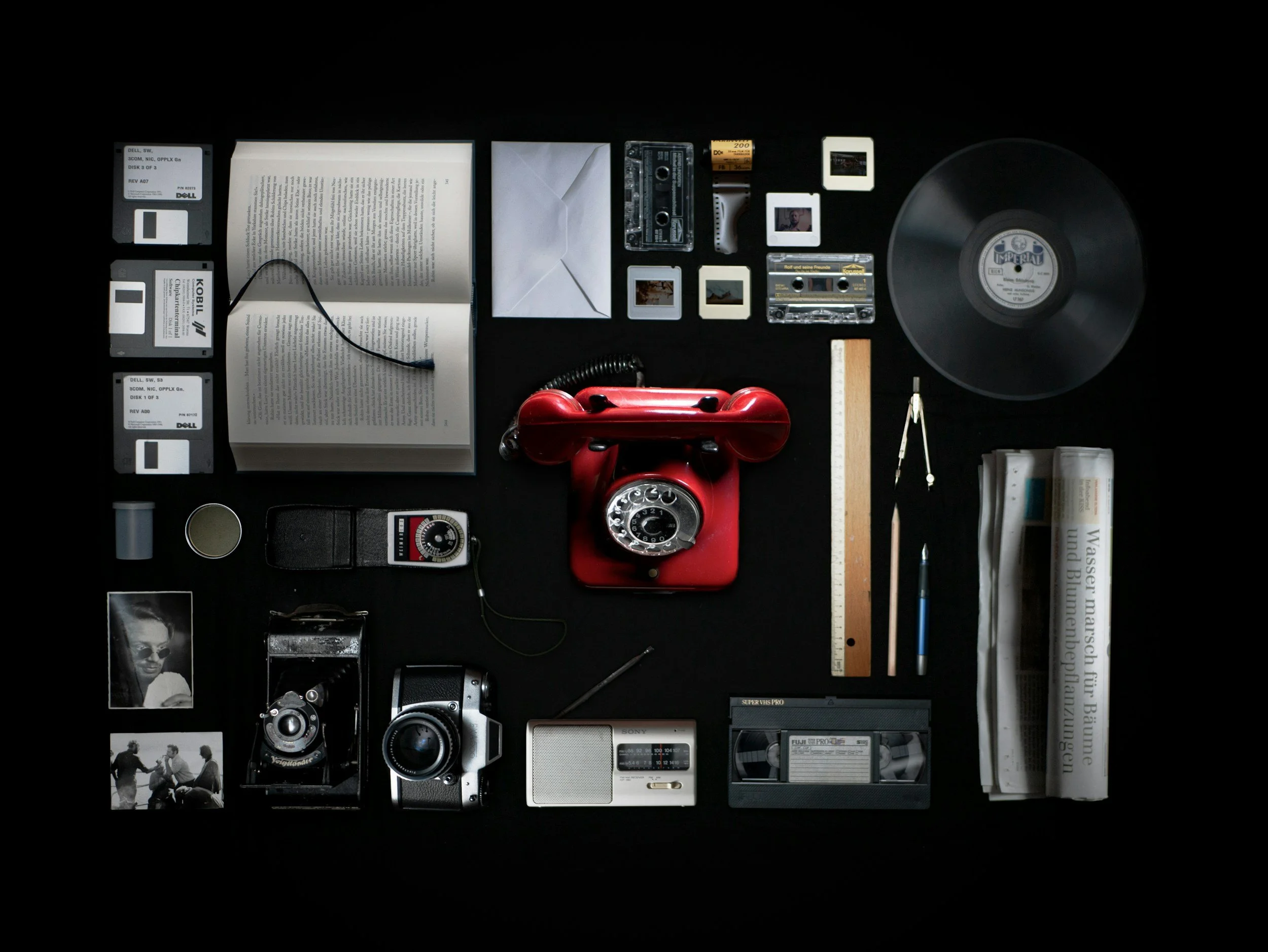 Verschiedene Gegenstände auf einer schwarzen Oberfläche, darunter eine rote Schneckenhaube, Kassetten, Fotografien, eine Uhr, ein Telefon, ein Radiergummi, ein Lineal, ein Taschenrechner, eine Kamera und Bücher.