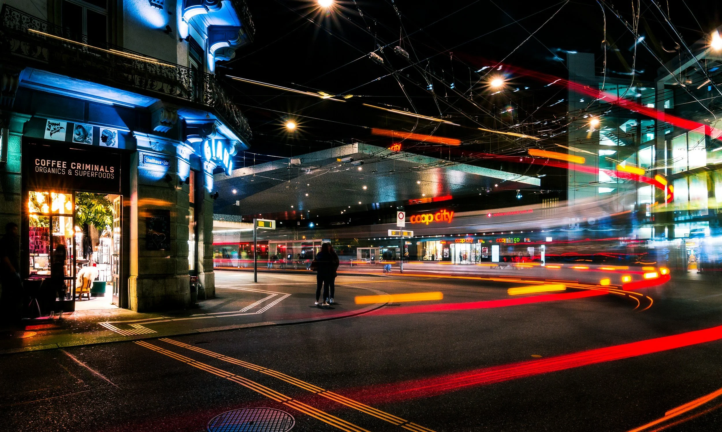 Lichtspuren von fahrenden Autos in einer Stadt bei Nacht, im Vordergrund ein Geschäft mit dem Schild 'Coffee Criminals' und mehreren Menschen auf der Straße.