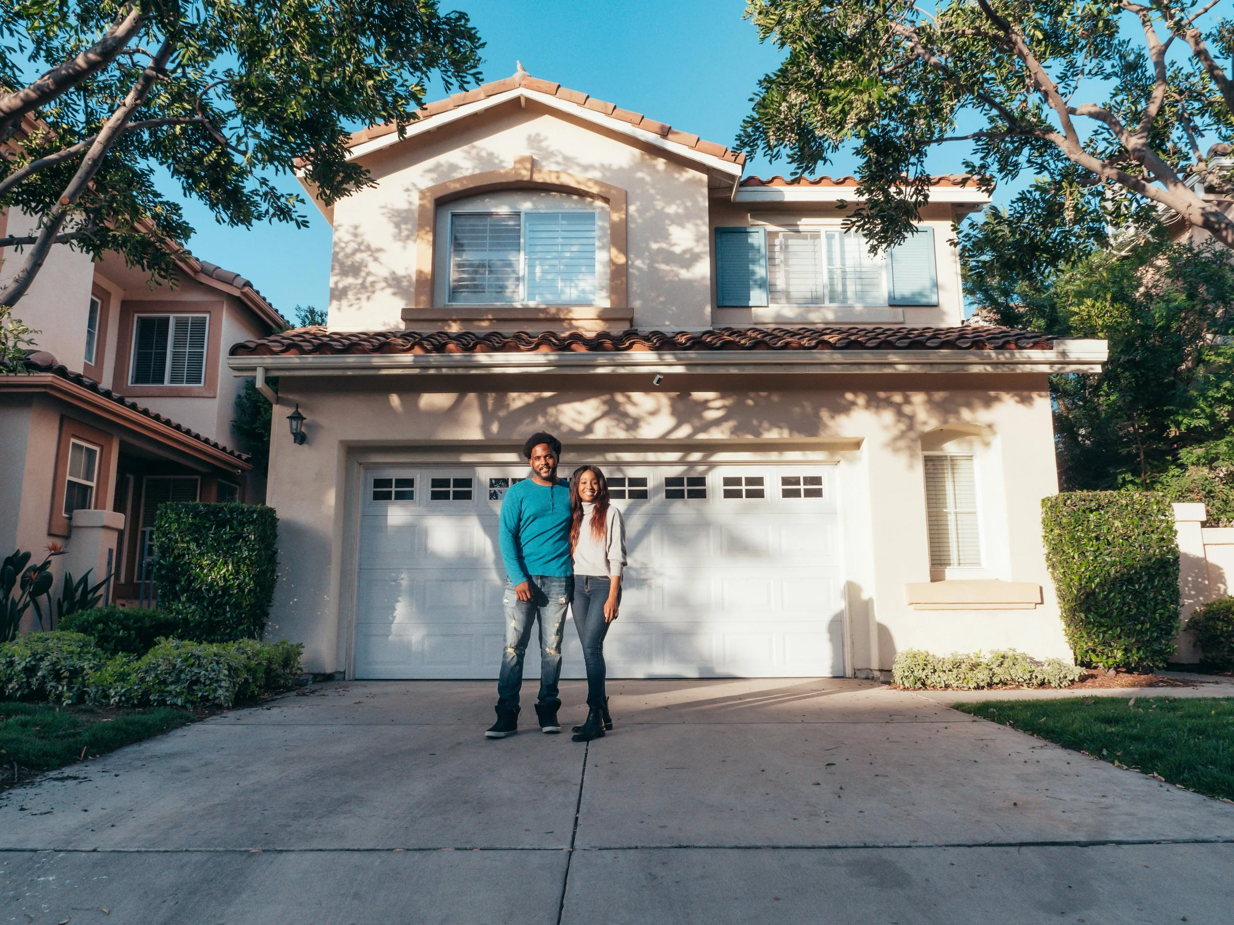 A happy young couple standing in their driveway in front of a beautiful two-story home in Forest Grove, Oregon, after following their 2026 real estate game plan.