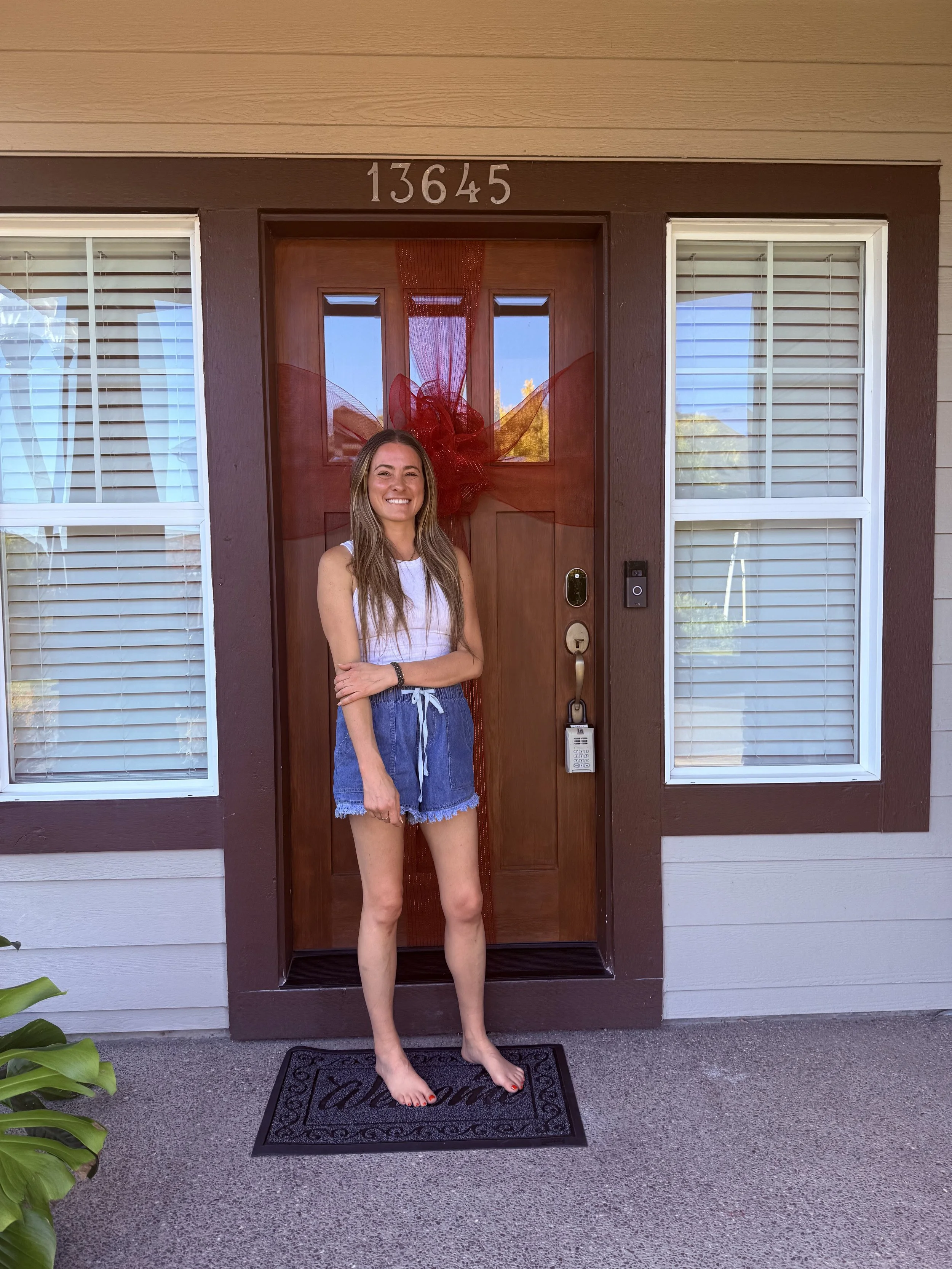 First-time homebuyer Brooke smiling on the front porch of her new home in King City, Oregon, standing in front of a wooden door decorated with a large red ribbon bow.