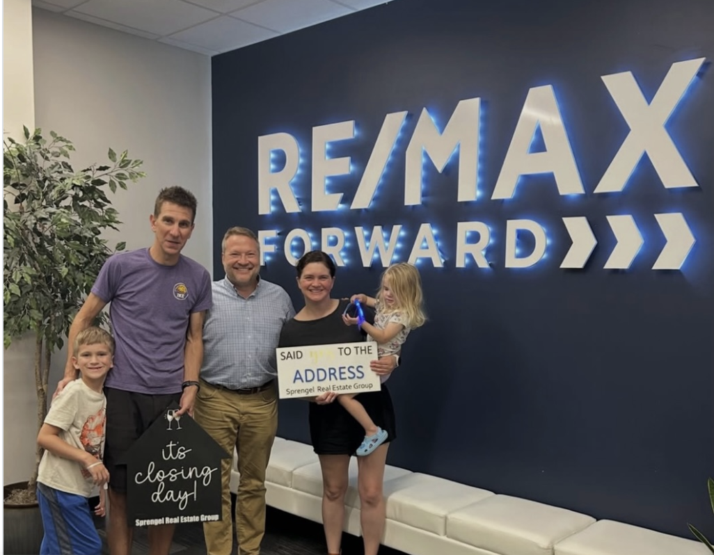 Happy family celebrating Closing Day at RE/MAX Forward office in Wisconsin holding signs reading 'It's closing day!' and 'Said YES to the ADDRESS'.