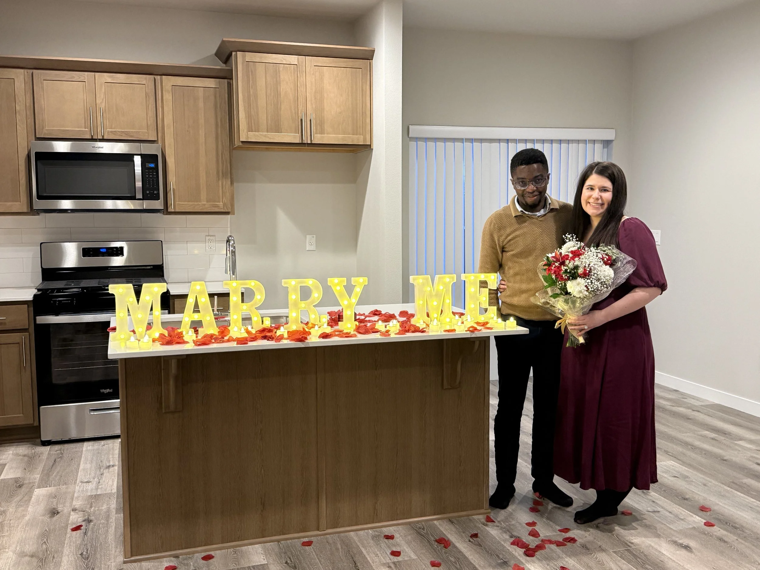 A couple standing in a kitchen with a 'Marry Me' sign illuminated on a counter decorated with red rose petals. The woman holds a bouquet of flowers, and they are smiling.