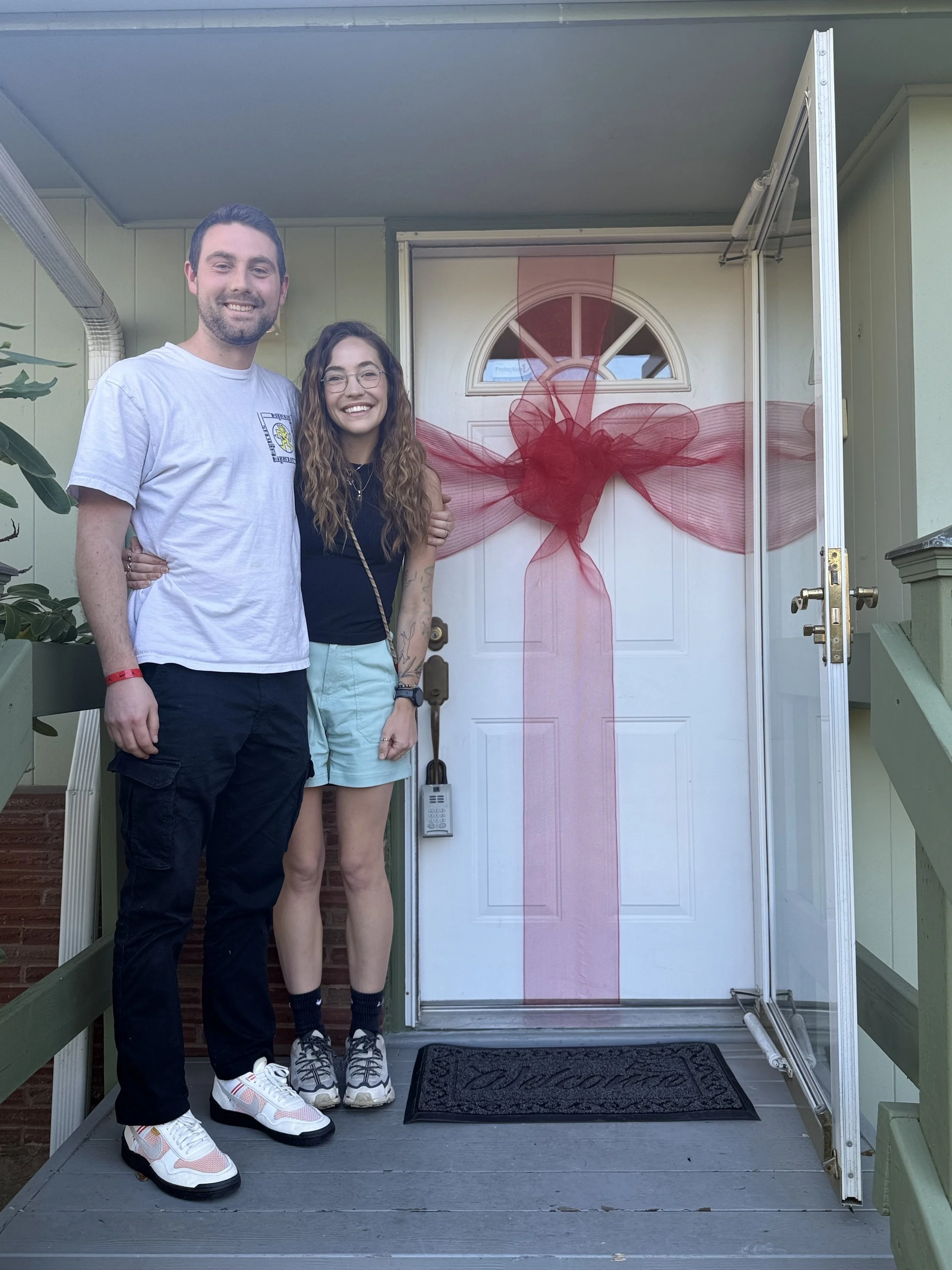 First-time home buyers Suze and Trey smiling happily together on the porch of their new home in Milwaukie, Oregon, standing in front of a white front door decorated with a large red ribbon bow.