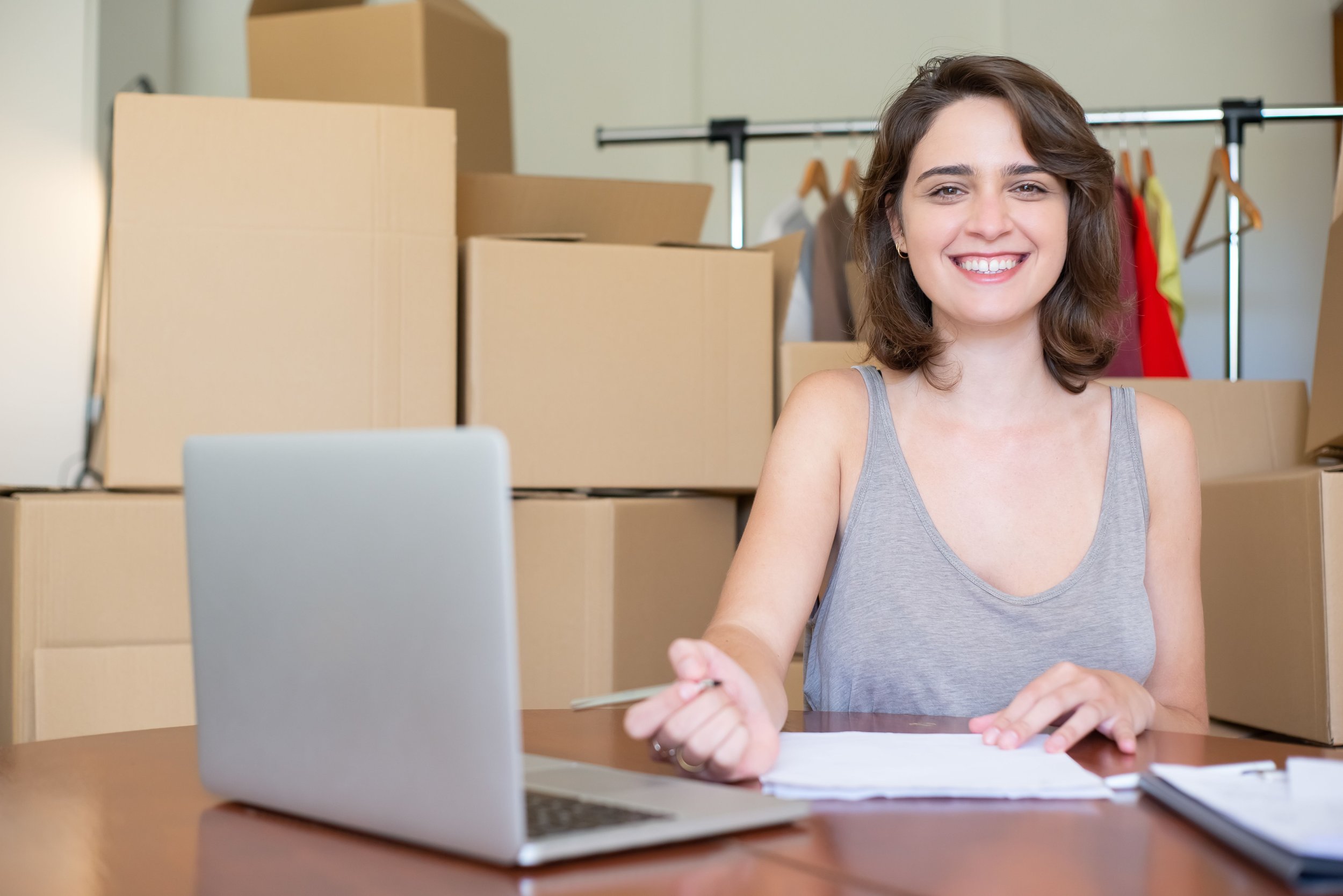 Smiling woman organizing moving plans and documents on a laptop surrounded by cardboard boxes in Beaverton Oregon