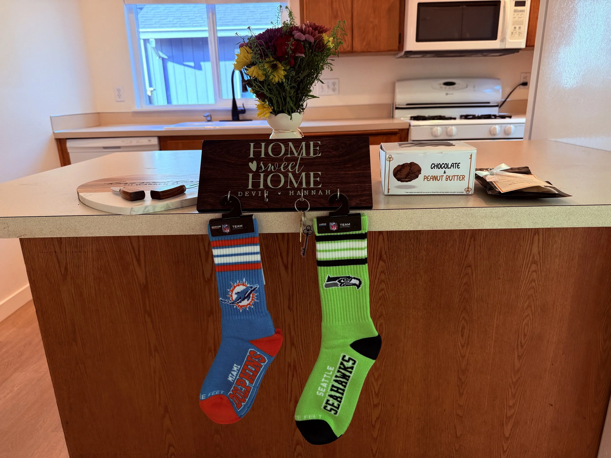 Personalized wooden key holder sign reading 'Home Sweet Home Devin and Hannah' displayed on a kitchen counter next to Miami Dolphins and Seattle Seahawks socks, cookies, and fresh flowers.