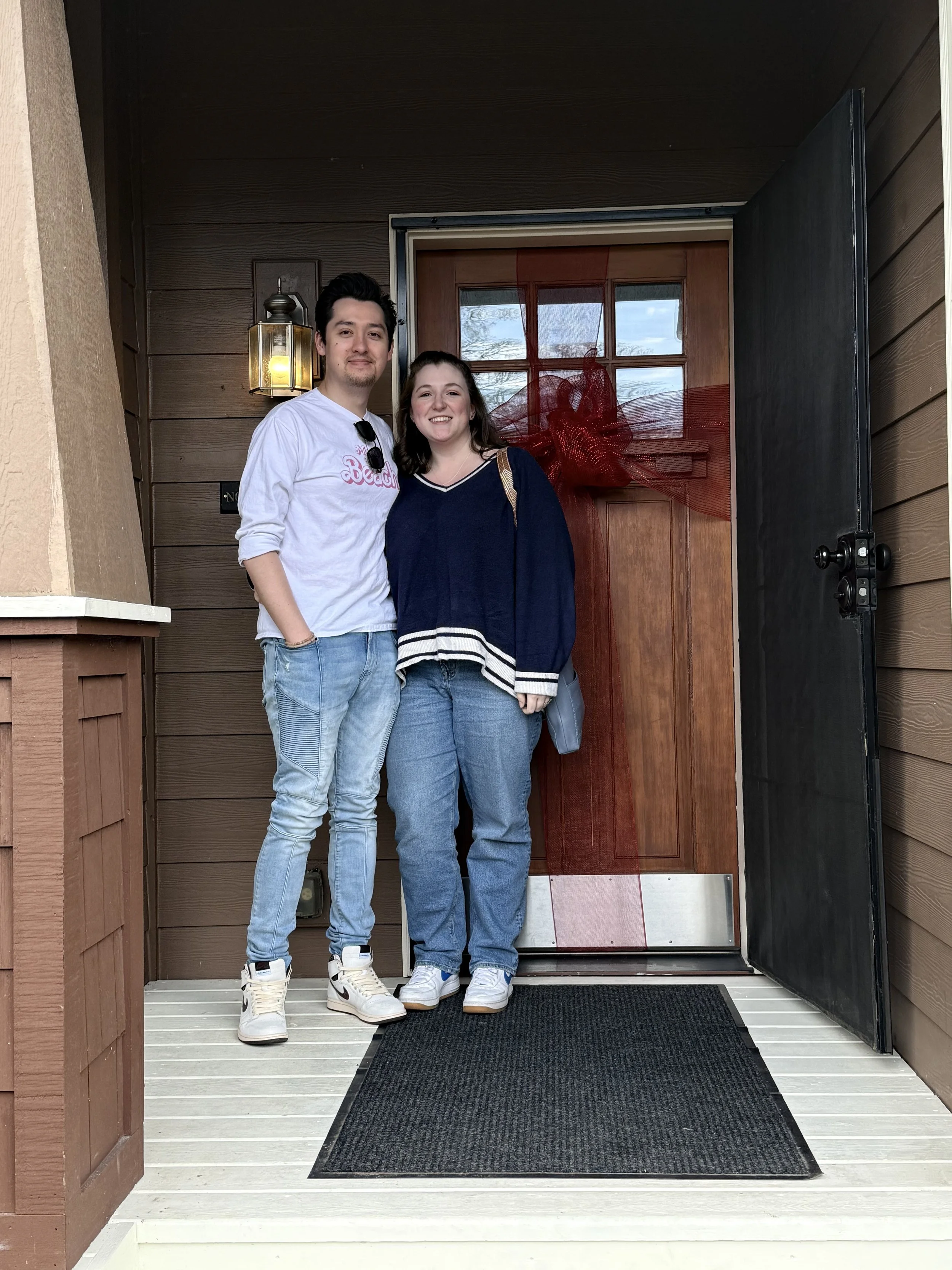 Happy new home buyers Harrison and Nicole smiling together on the porch of their new house in Portland, standing in front of a wooden front door decorated with a large red bow.
