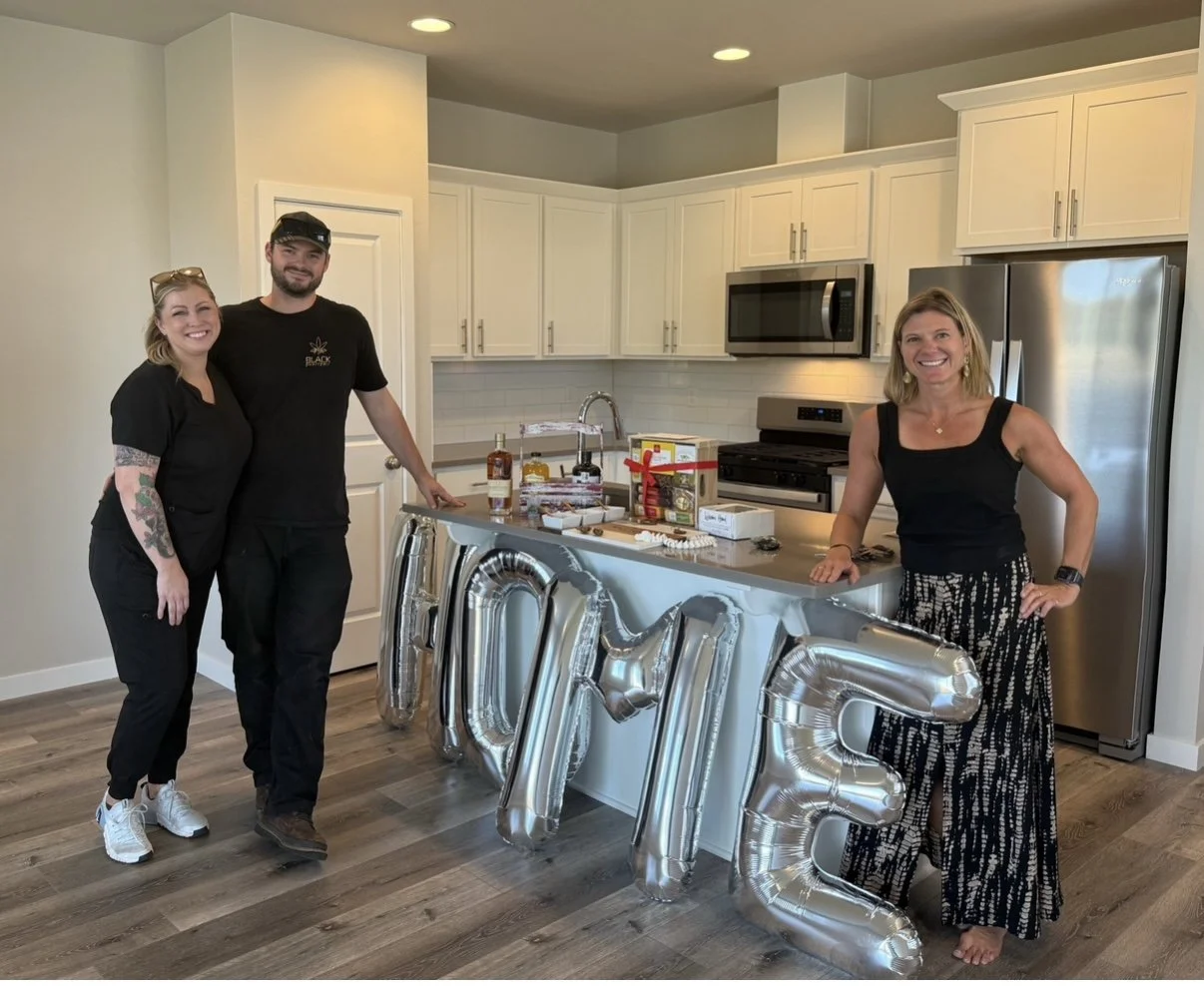 Three people smiling in a kitchen next to a silver 'LOVE' balloon arrangement and a counter with gifts and drinks, with white cabinets and stainless steel appliances in the background.