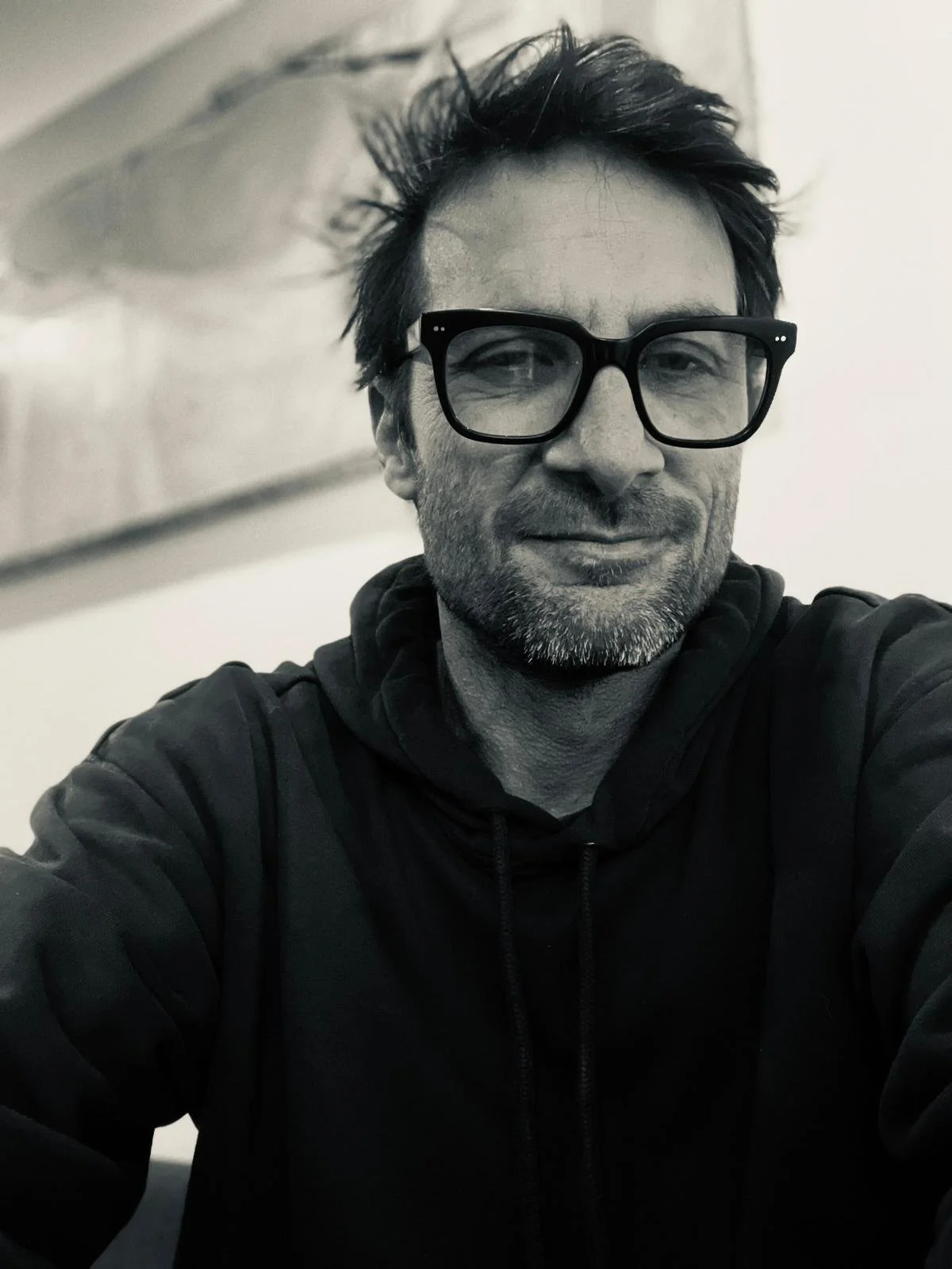 A black and white close-up selfie of a man with messy hair, wearing large glasses and a hooded sweatshirt, smiling slightly.