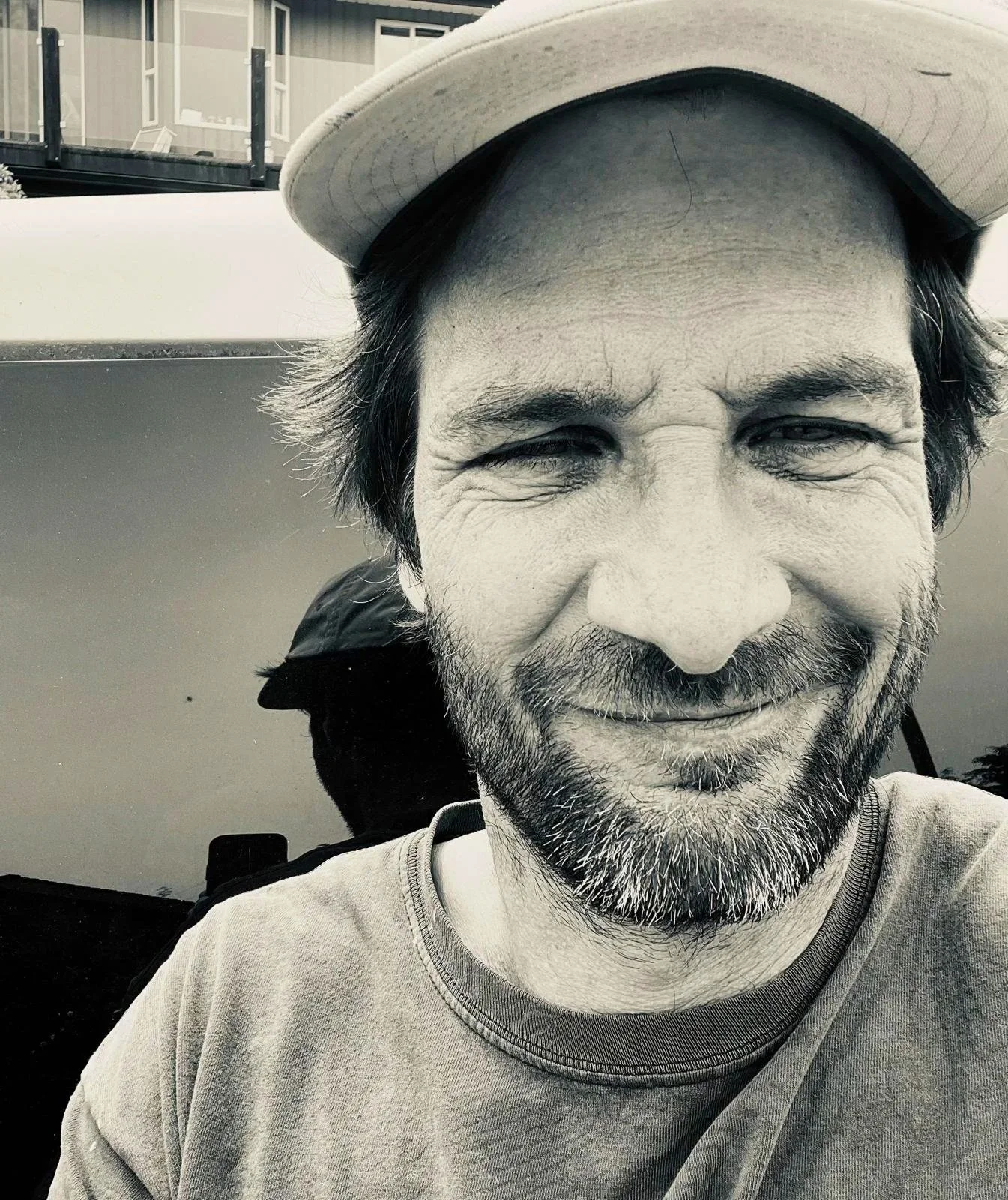 Close-up black and white photo of a smiling man with a beard and mustache, wearing a baseball cap and a t-shirt, outdoors near a building and a boat.
