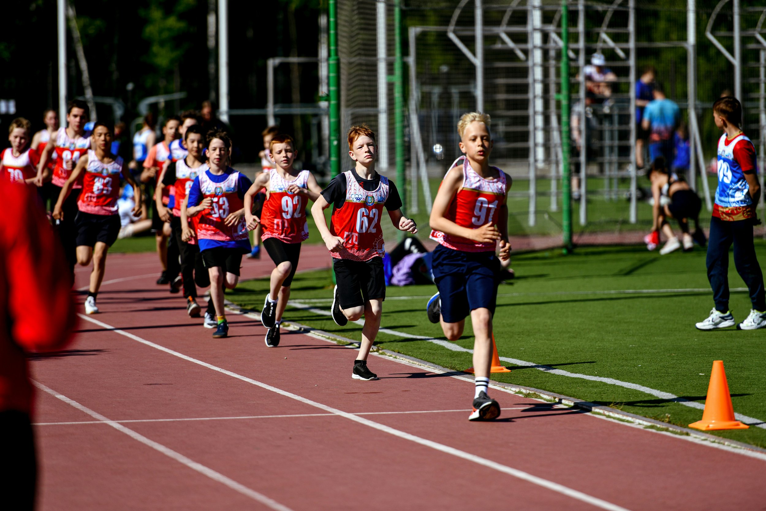 Young children participating in a track race on an outdoor running track, wearing sports uniforms and running near orange cones.