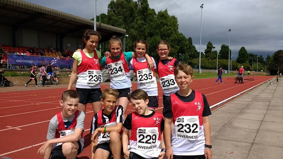Group of young children at a track and field event, wearing matching red and black athletic uniforms and numbered bibs with 'Inverness' on them.