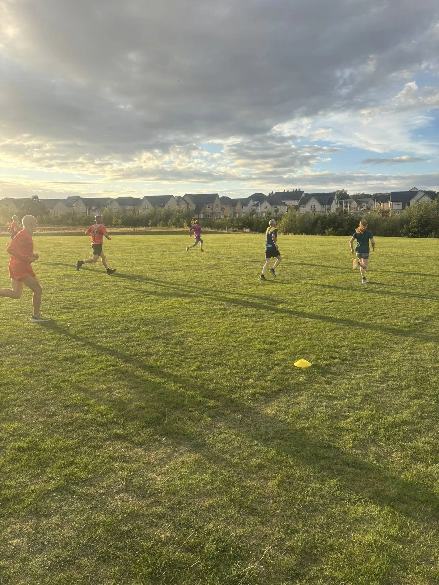 Club members  running on a grassy field doing an interval session, with houses and a cloudy sky in the background.