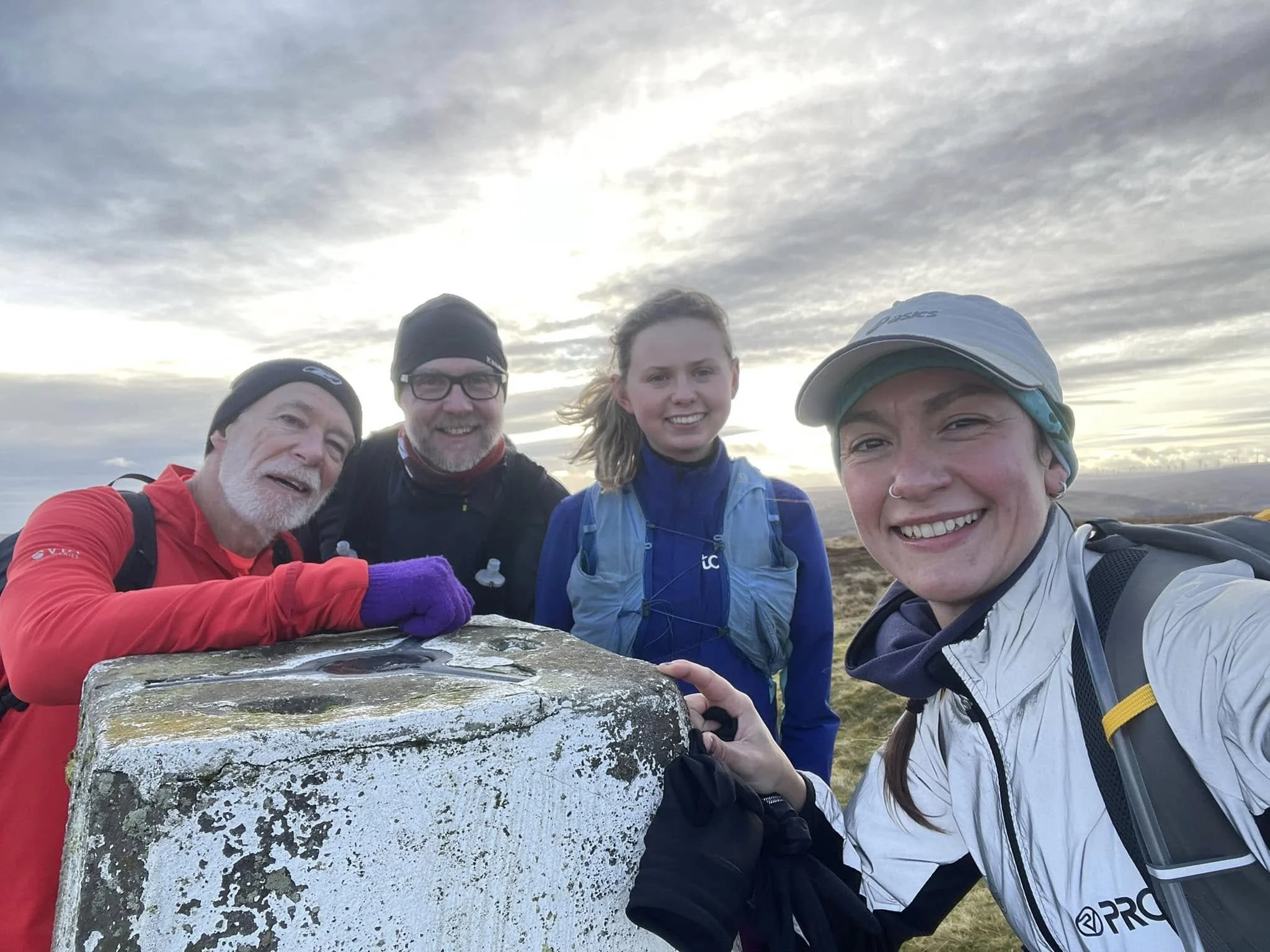Club members smiling outdoors on a cloudy day, with a young woman in the front taking a selfie; they are standing beside a trig pillar in a natural landscape.