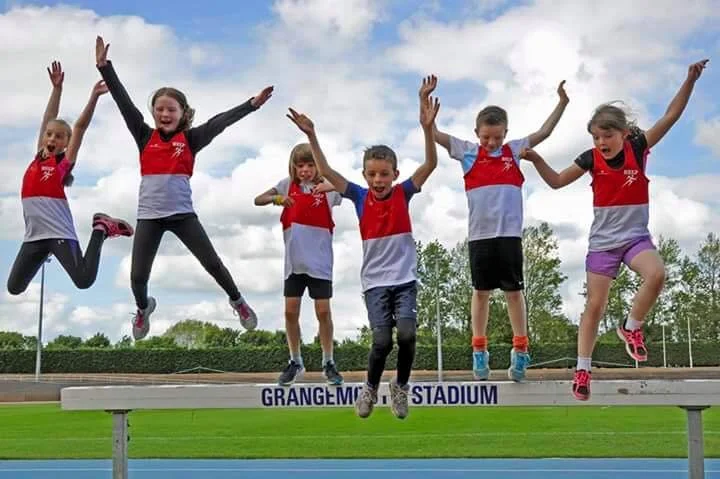 Six children jumping together on a running track at Grange-Melstad Stadium.