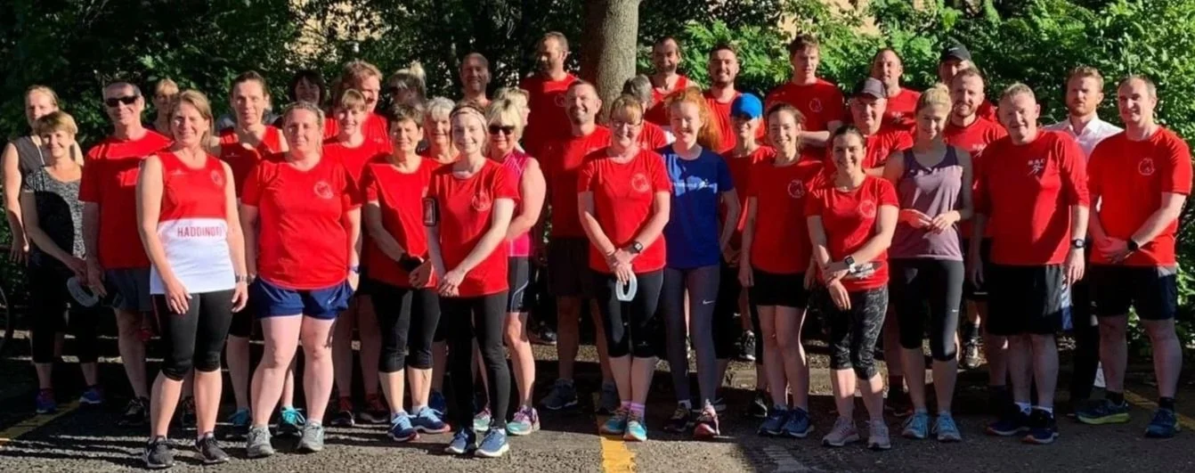 Group photo of members of Haddington Running Club in the club red t-shirt, standing outdoors on a sunny day, in front of green trees.