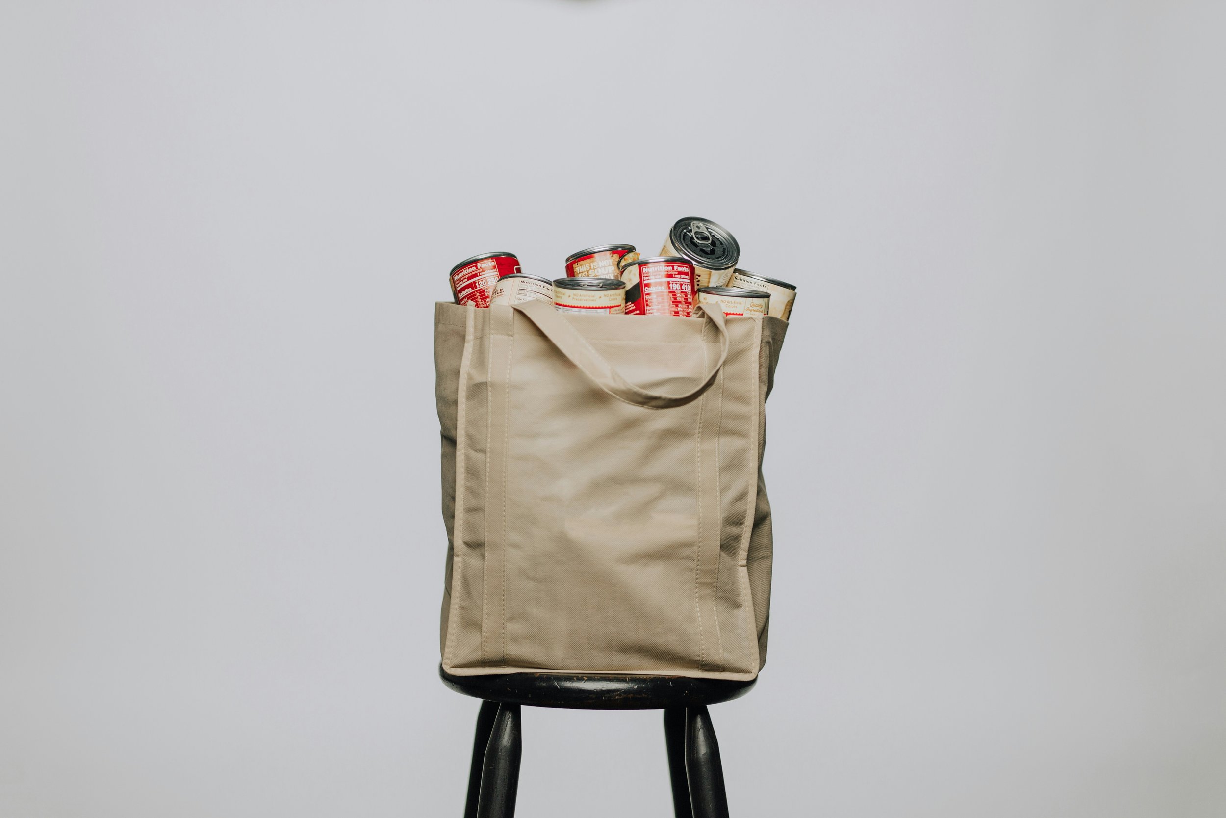 Beige reusable canvas tote bag filled with canned food items, placed on a black stool against a plain white background.