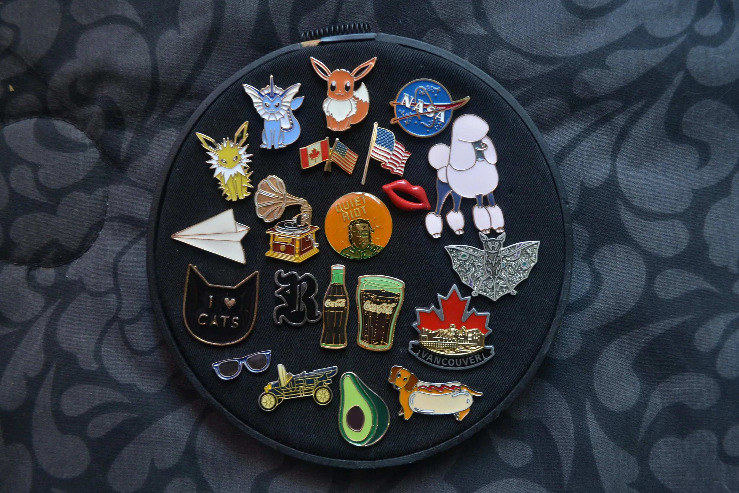 Collection of colorful enamel lapel pins displayed on a round black board, featuring pop culture icons, flags, animals, and graphic designs.