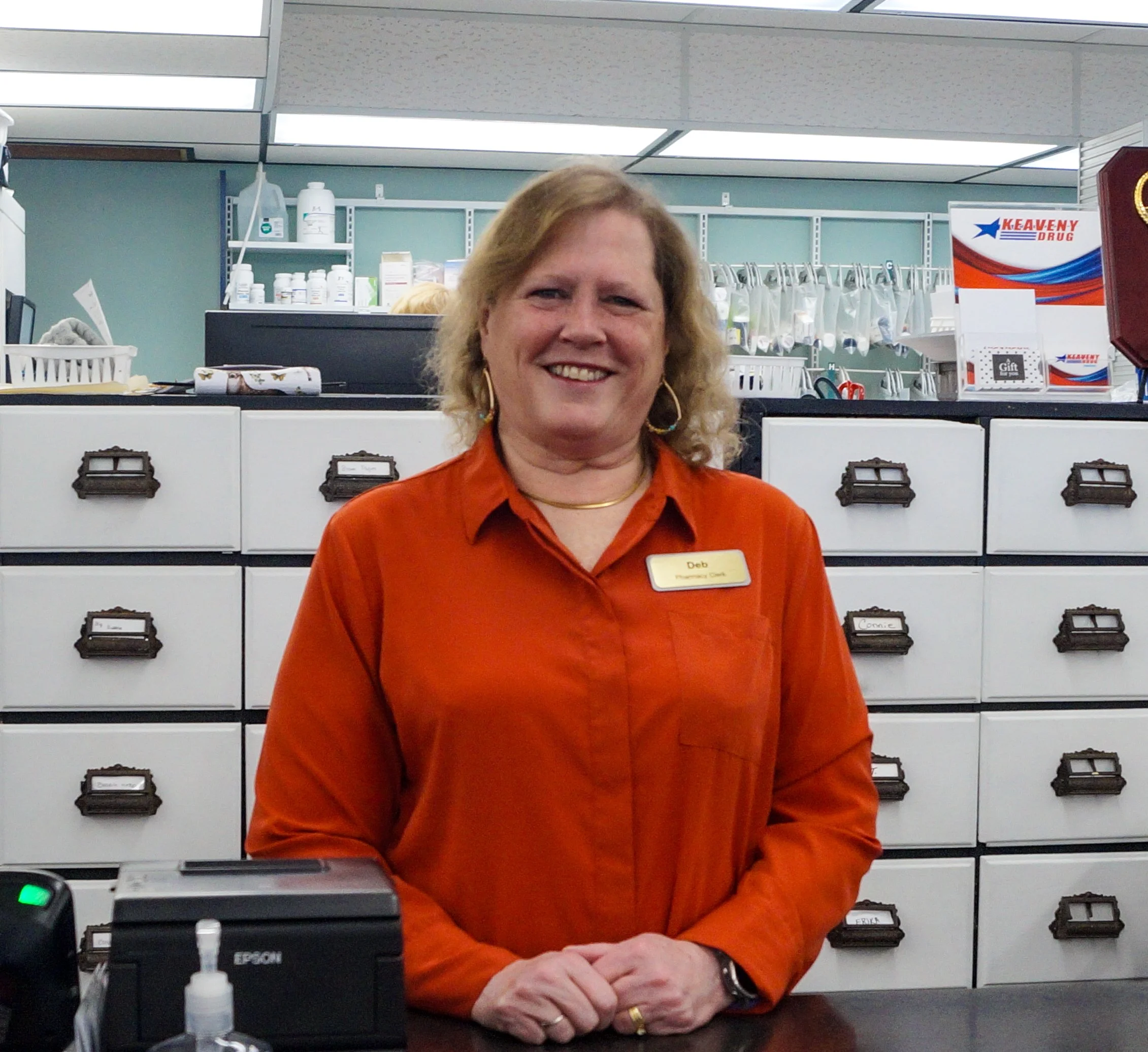 A woman in an orange shirt with a name tag stands behind a counter inside a pharmacy or medical office, smiling at the camera.