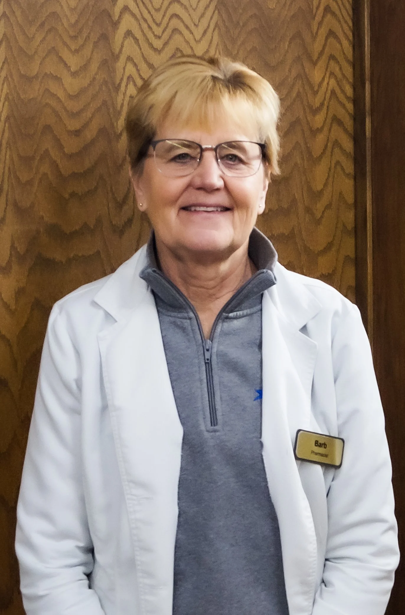 A woman wearing glasses, a white coat, and a name tag that reads 'Barb', standing in front of a wood-paneled wall, smiling at the camera.
