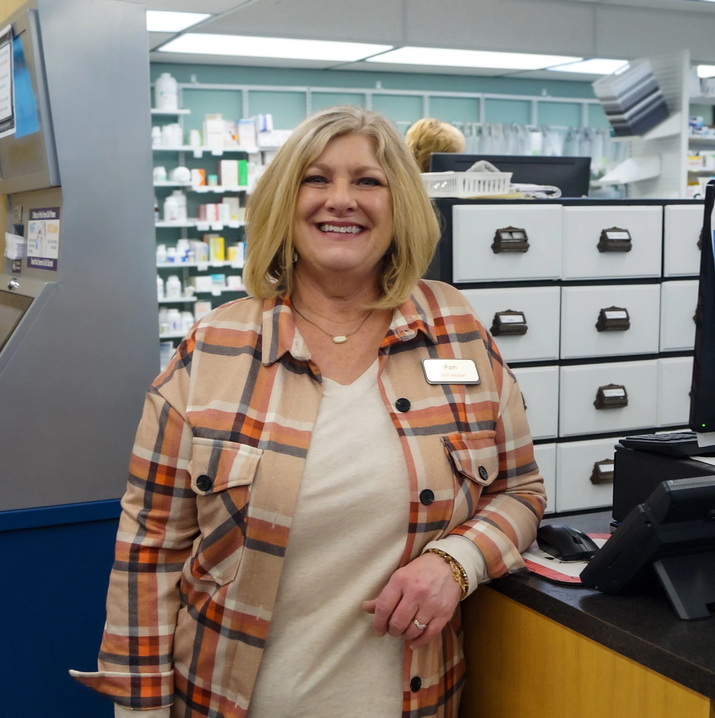 A smiling woman wearing a plaid shirt with a name tag standing at a pharmacy counter.