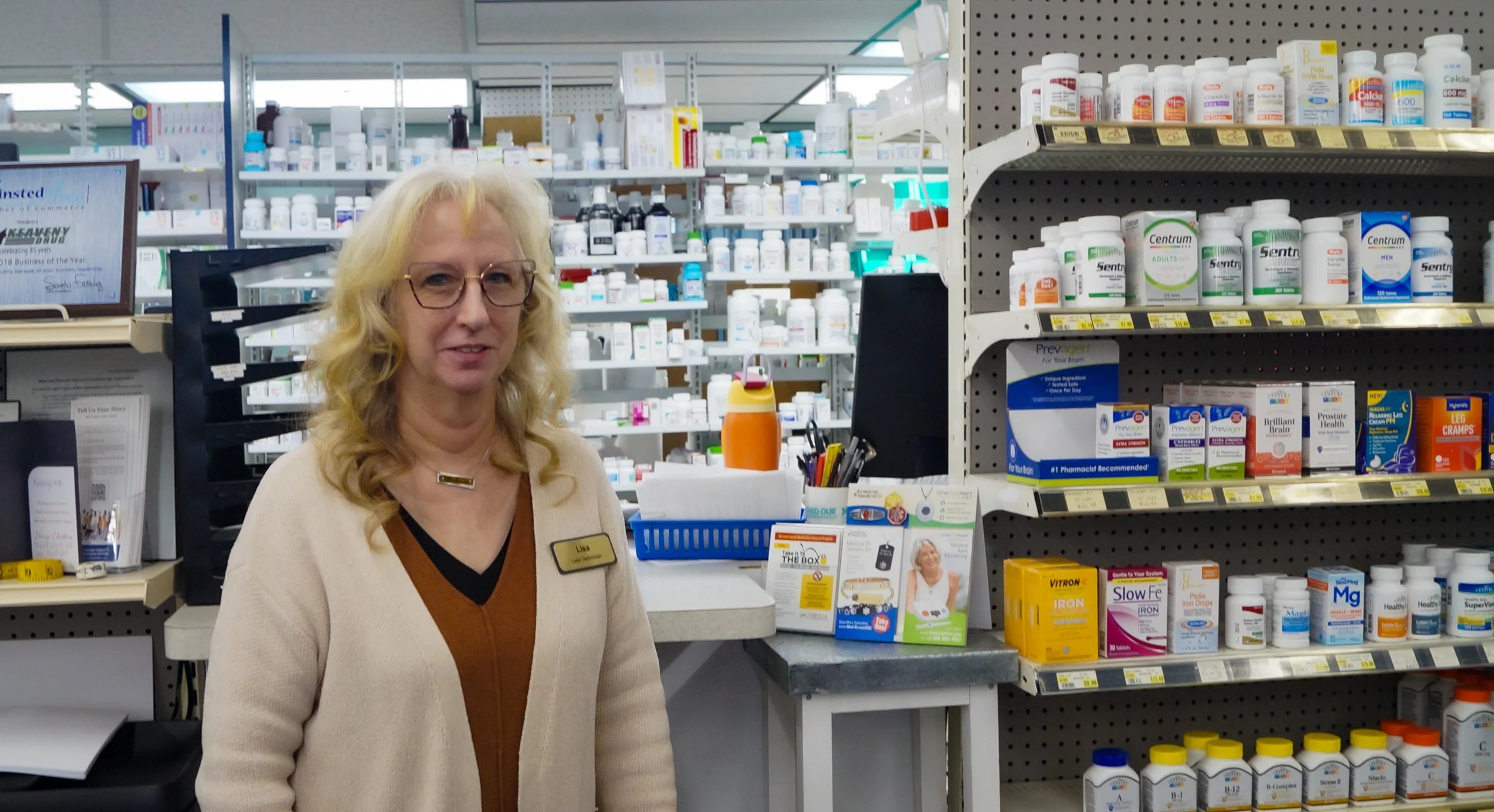 A woman with blonde hair, glasses, and a registration badge standing in front of pharmacy shelves stocked with vitamins and supplements.