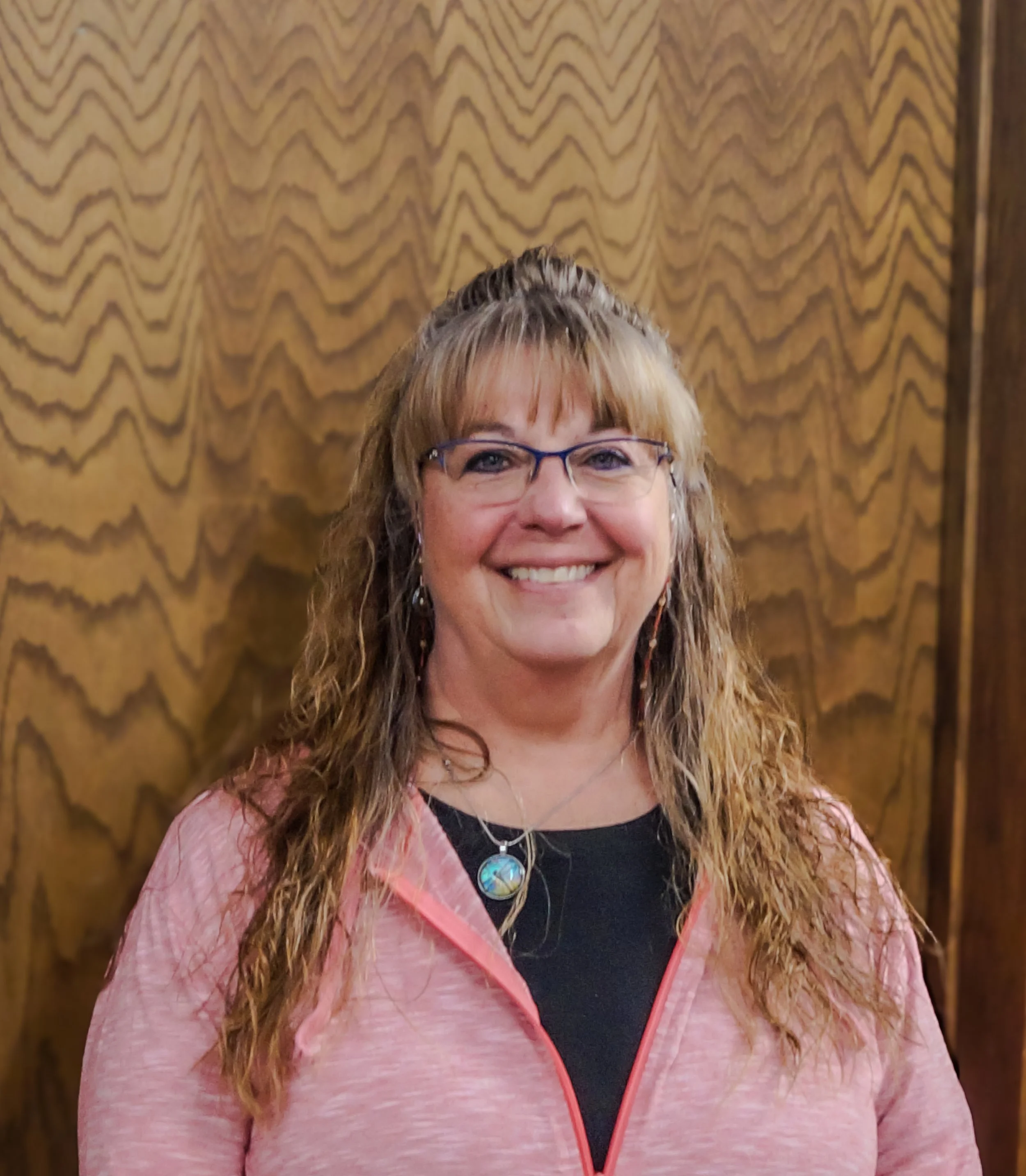 A woman with curly blonde hair, glasses, and earrings, smiling at the camera. She is wearing a pink jacket over a black top and a necklace with a round pendant. The background features a wood-paneled wall.