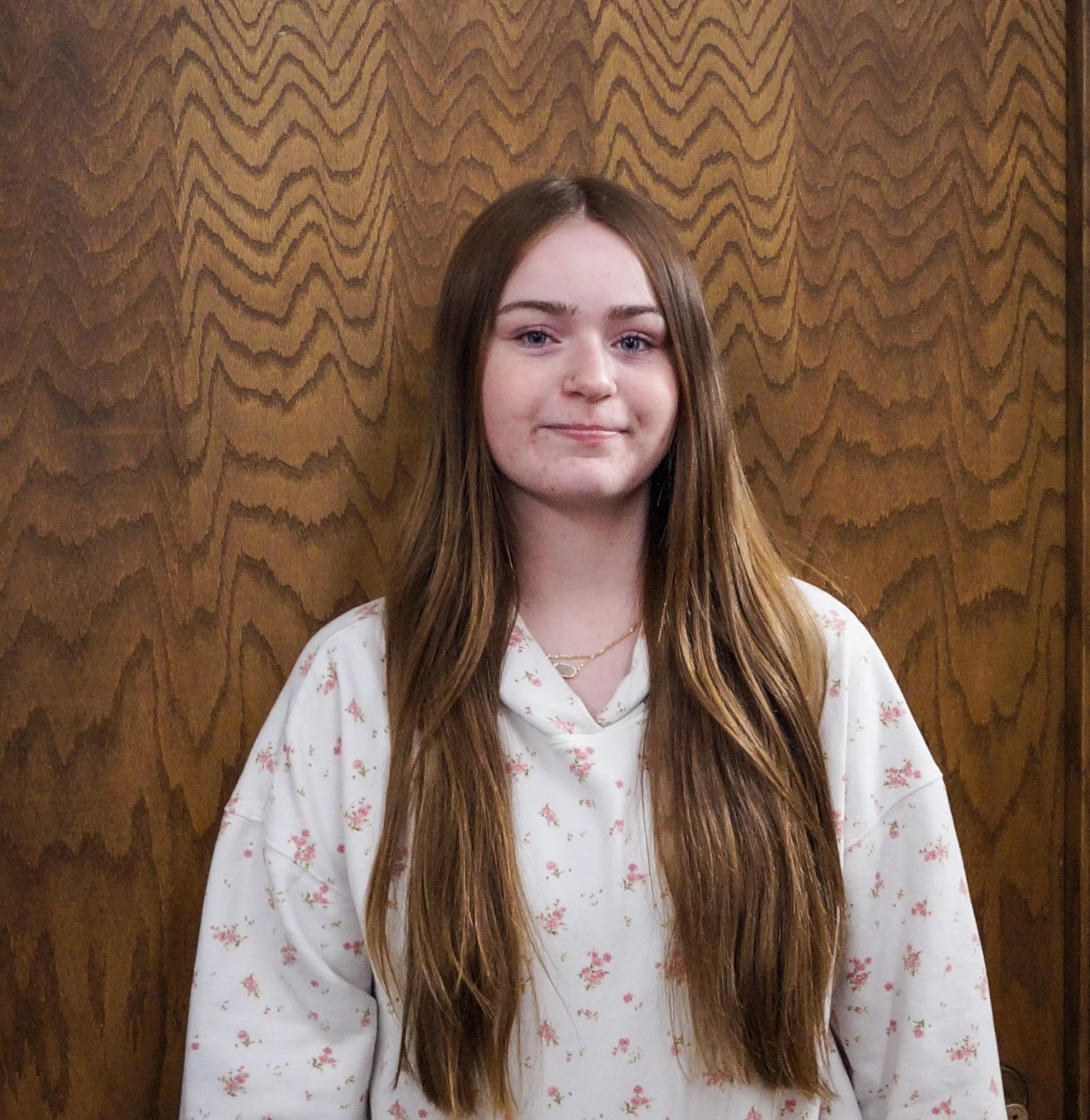 Young woman with long brown hair wearing a white floral shirt standing in front of a wooden wall.