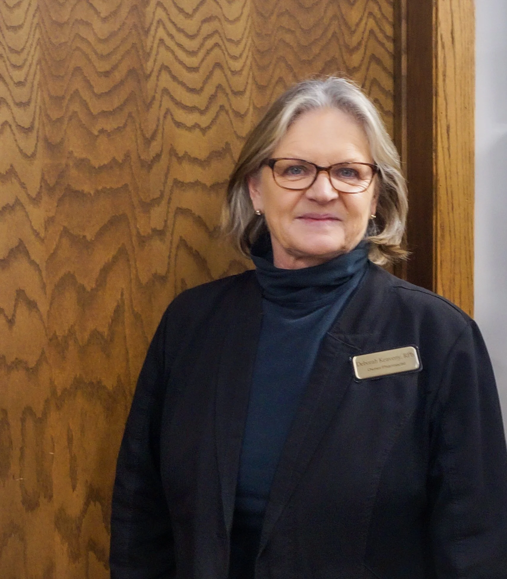 A woman with glasses and short, gray hair standing indoors against a wood-paneled wall. She is wearing a dark blazer and a navy scarf, with a name tag that reads 'Deborah Keaveny'.