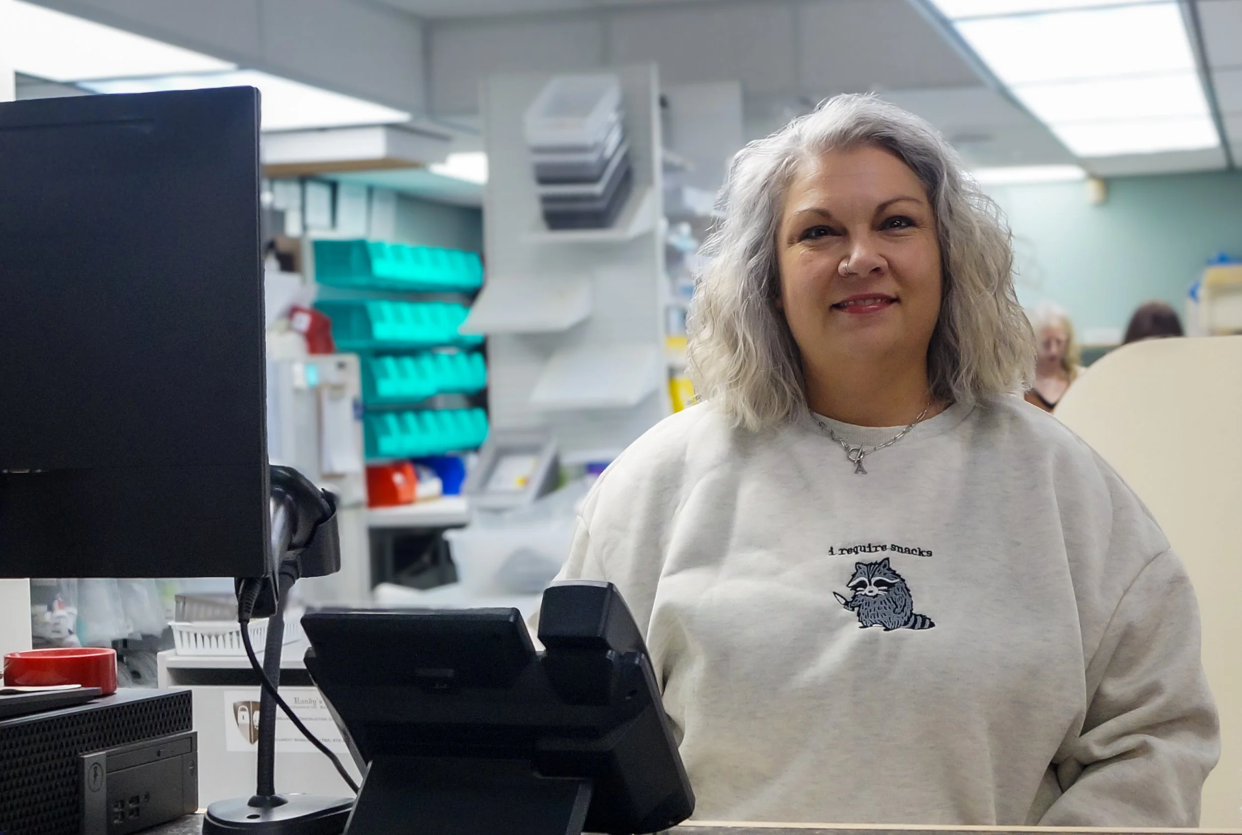 A woman with gray curly hair smiling behind a checkout desk in a store, wearing a sweatshirt with a raccoon illustration and the text 'I require snacks'.