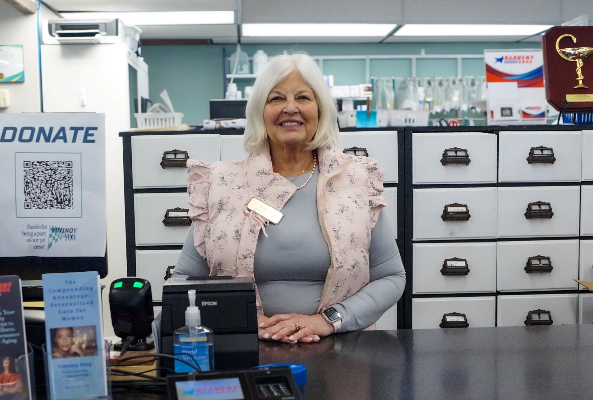 A smiling elderly woman with white hair, wearing a pink floral vest over a gray top, standing at a reception desk in a medical or office setting.