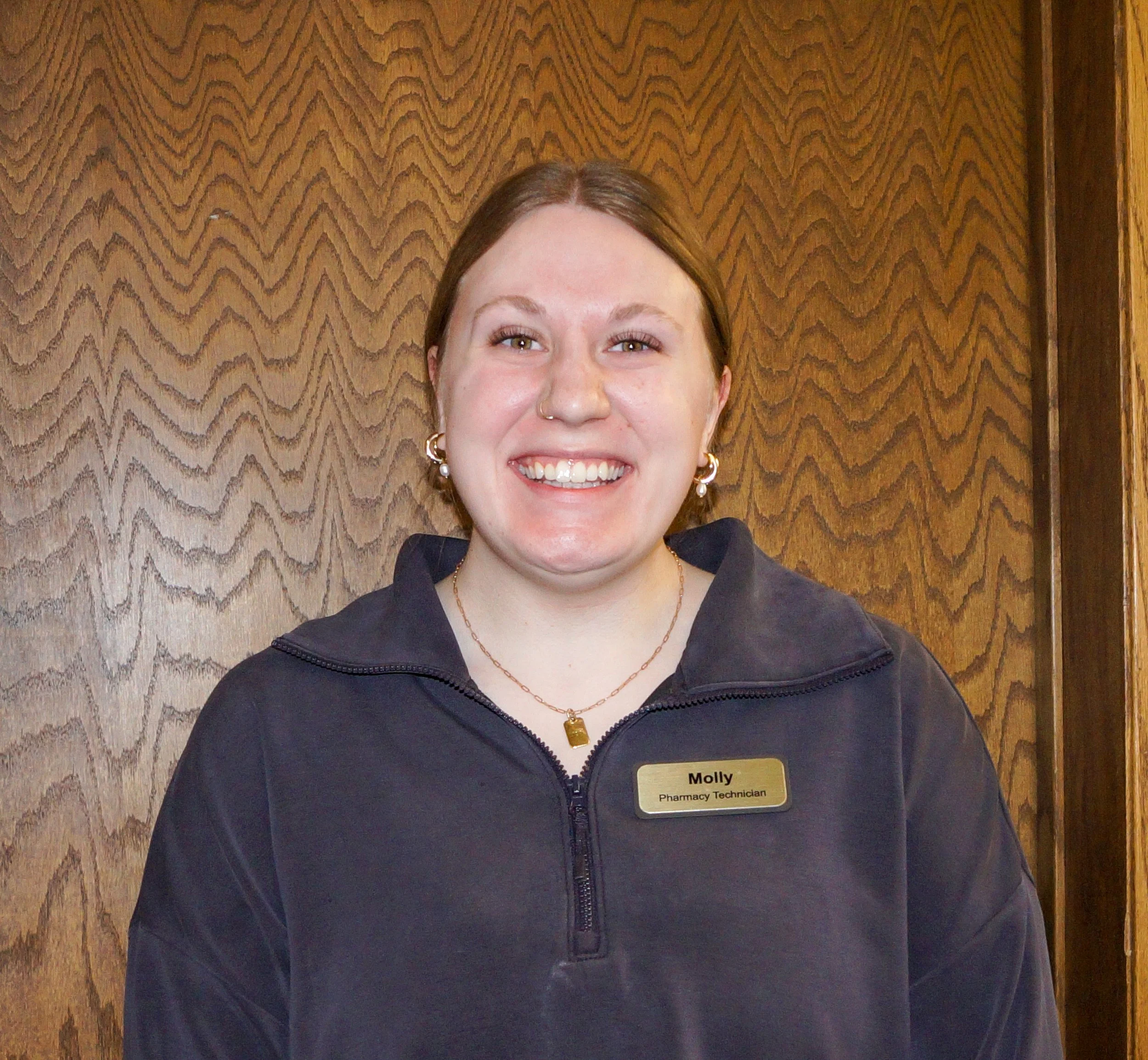 A smiling woman with short brown hair, glasses, wearing a beige sweater, standing behind a pharmacy counter, with a telephone and computer in front of her, and pharmacy shelves in the background.