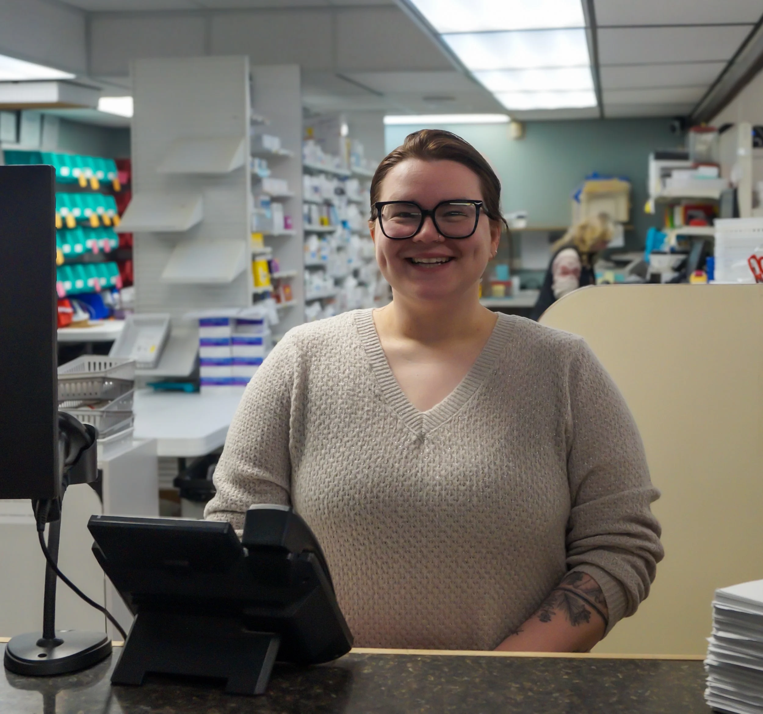 A smiling woman with short brown hair, glasses, wearing a beige sweater, standing behind a pharmacy counter, with a telephone and computer in front of her, and pharmacy shelves in the background.