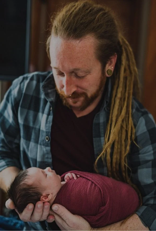 A man with long dreadlocks and beard gently holds a newborn wrapped in a burgundy blanket, looking down at the baby.